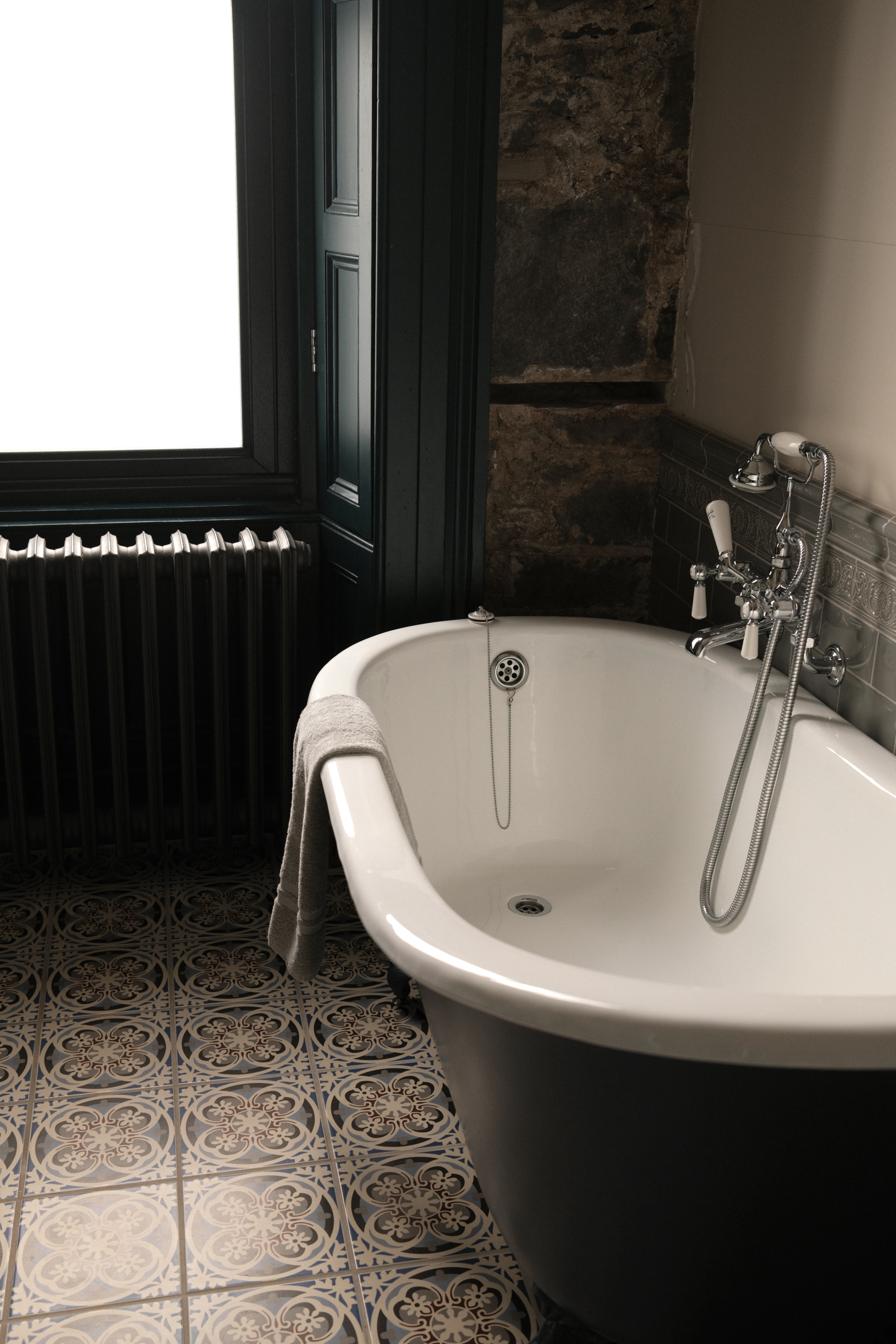 A roll-top bath with traditional taps stands against a stone wall, a folded towel on its rim and patterned tiles underfoot.