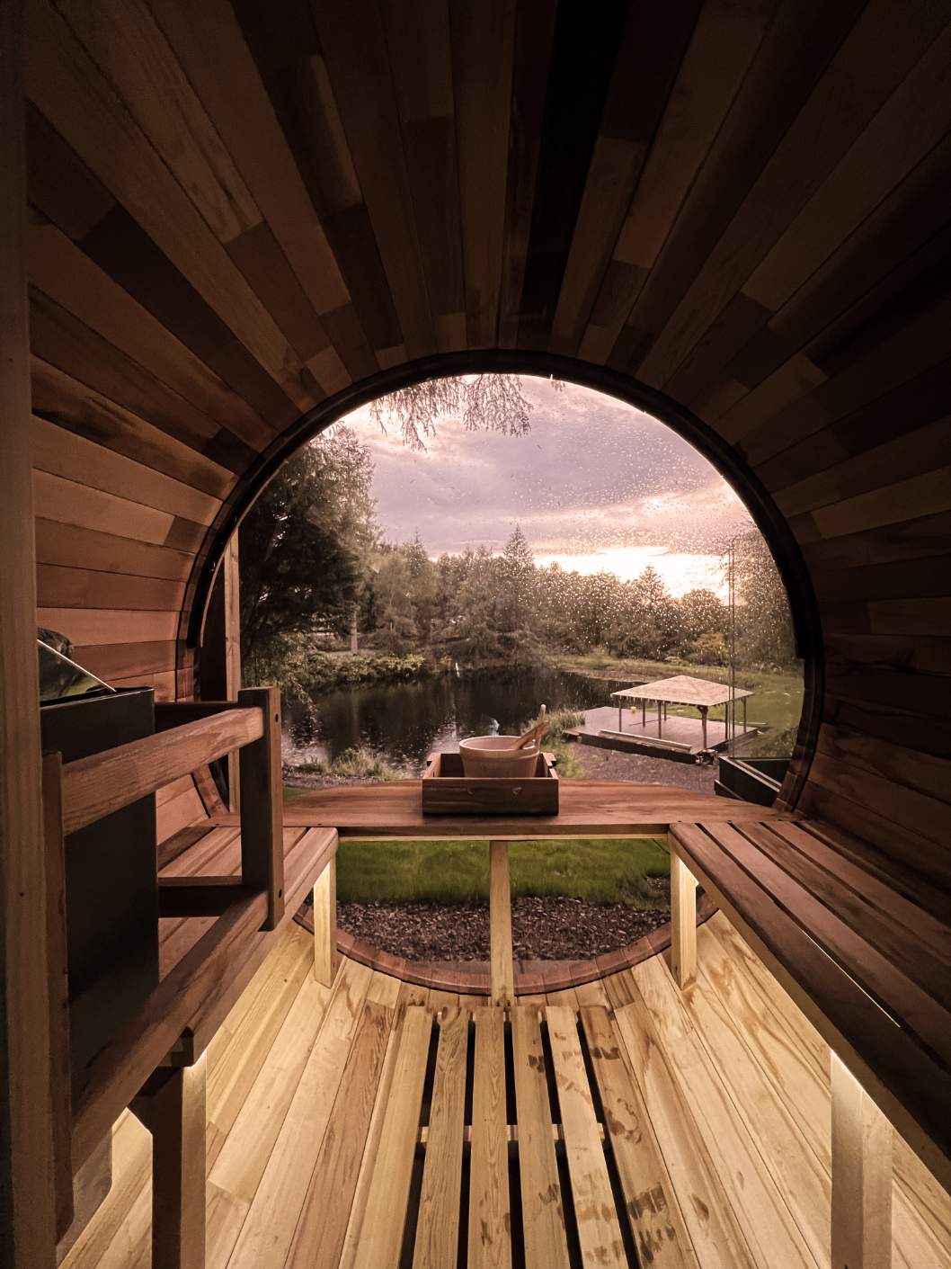 A curved timber sauna looks out through its round window onto a loch and a covered jetty at dusk, a small tray set on the deck outside.
