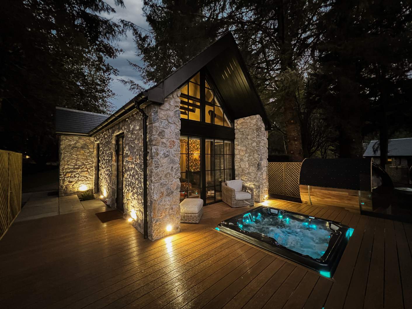 A stone and timber lodge glows at night, its gabled glazing lit above a hot tub set into the timber deck.