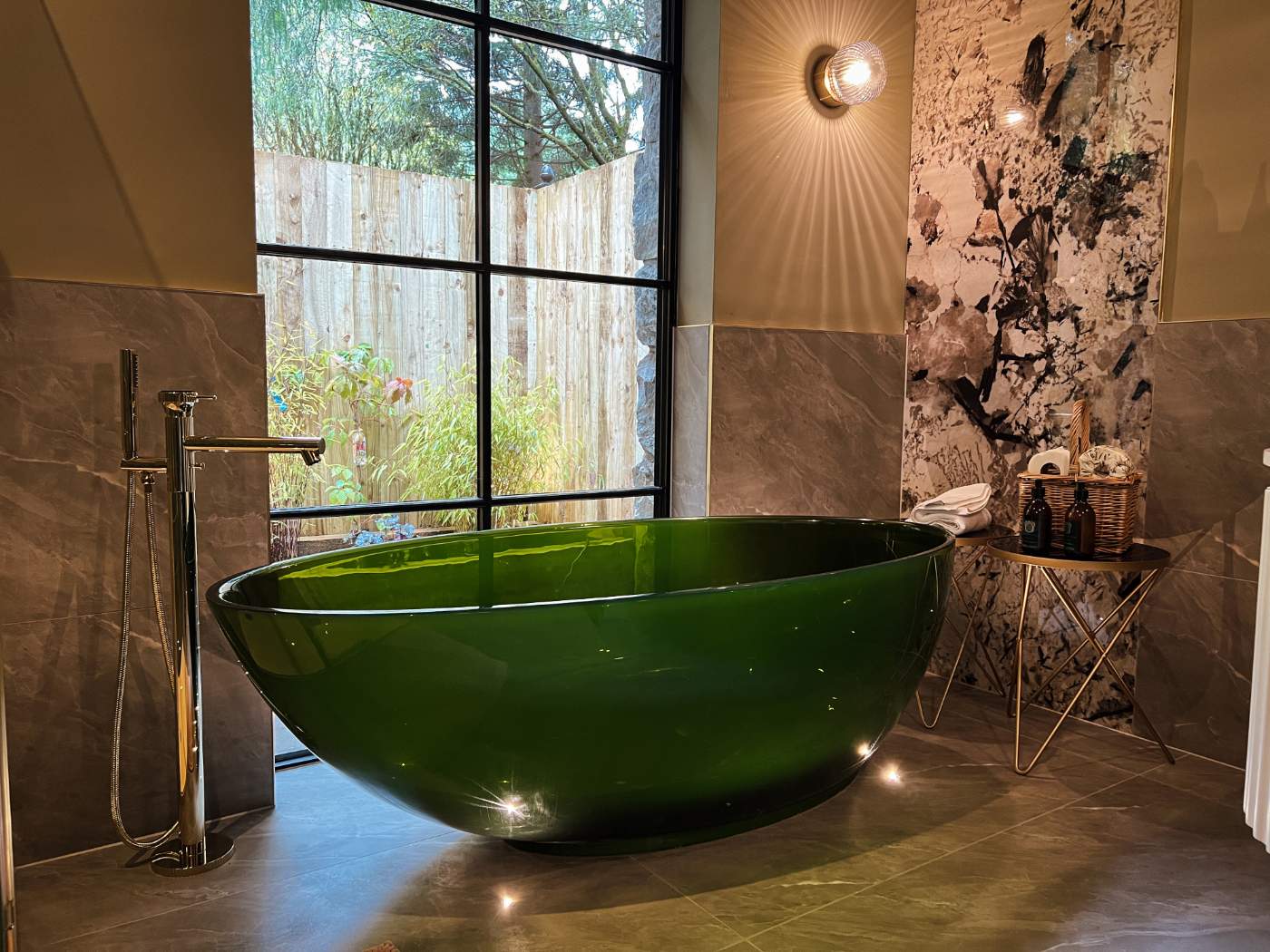 A deep green stone bath stands before a tall window, a chrome floor-standing tap beside it and folded towels on a side table.
