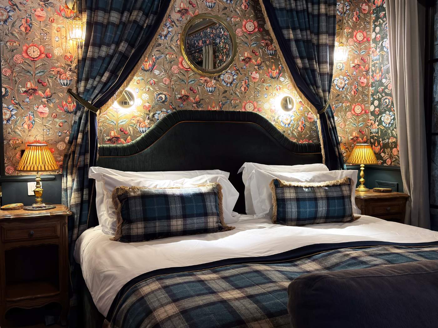 A bed with a dark upholstered headboard sits before wallpaper of birds and flowers, lit wall sconces either side and tartan cushions on the bedding.