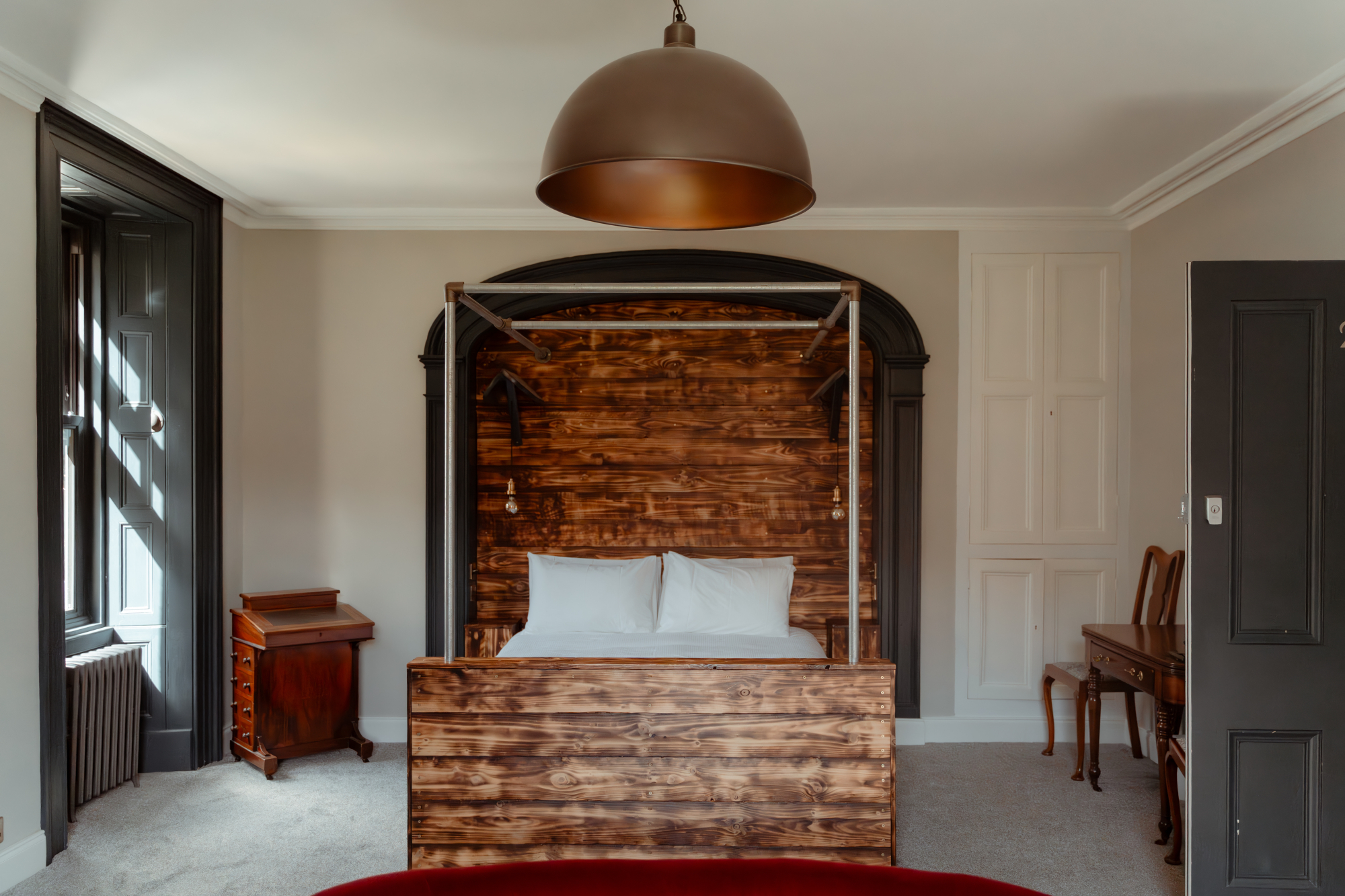 A four-poster bed with a scorched timber headboard sits beneath a copper dome light, an antique writing desk and piano visible through the archway behind.