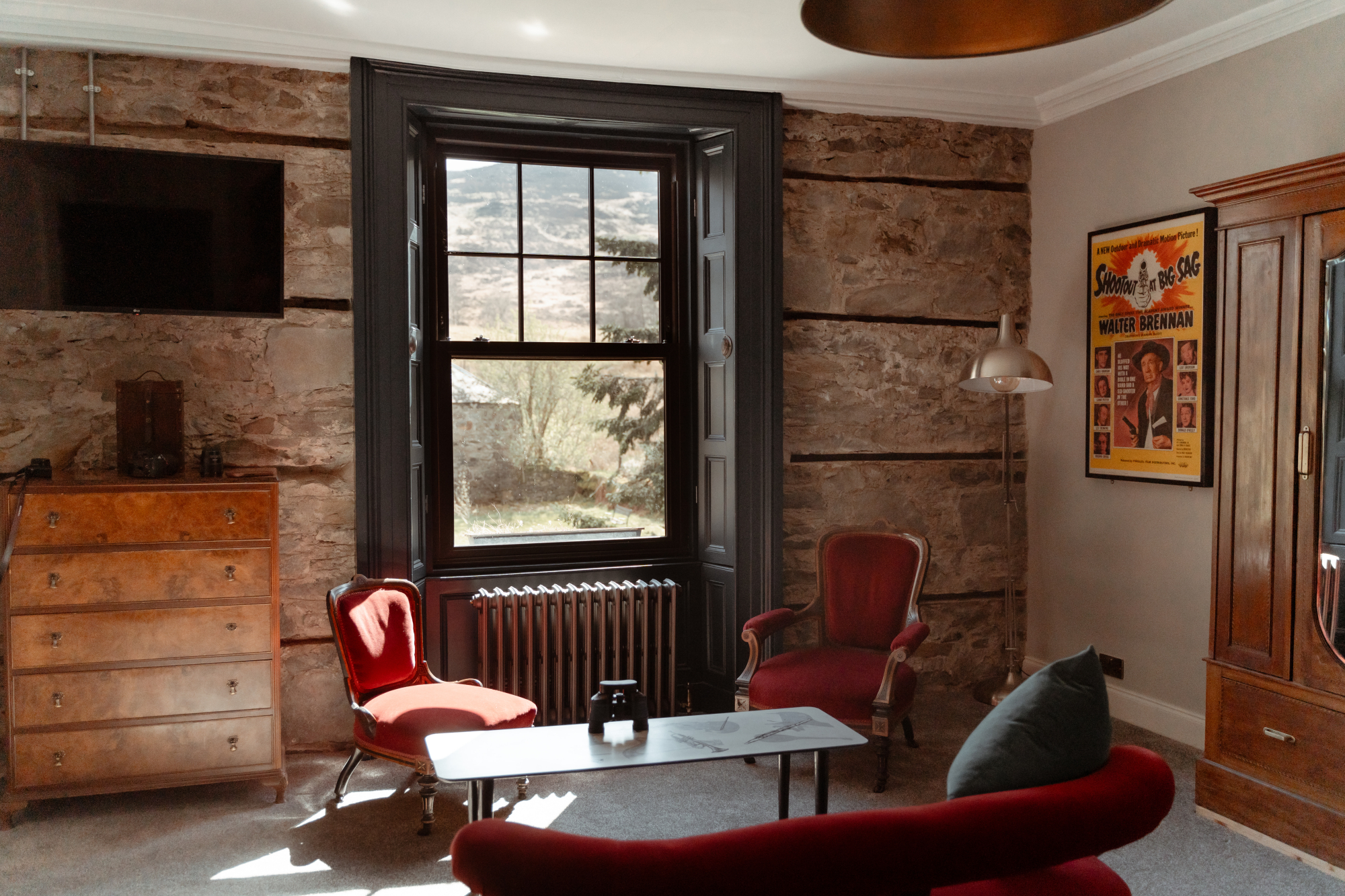 A red armchair and matching sofa sit before a stone wall hung with a vintage film poster, an antique wardrobe and a window framing hills to the side.