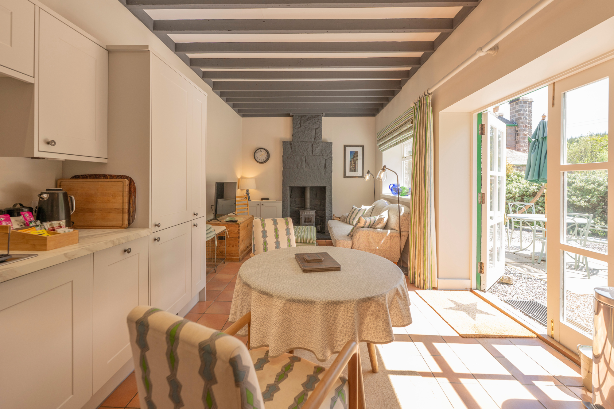A round table set beneath a beamed ceiling stands before a wood burning stove and sofa, glazed doors opening onto a patio with an umbrella.