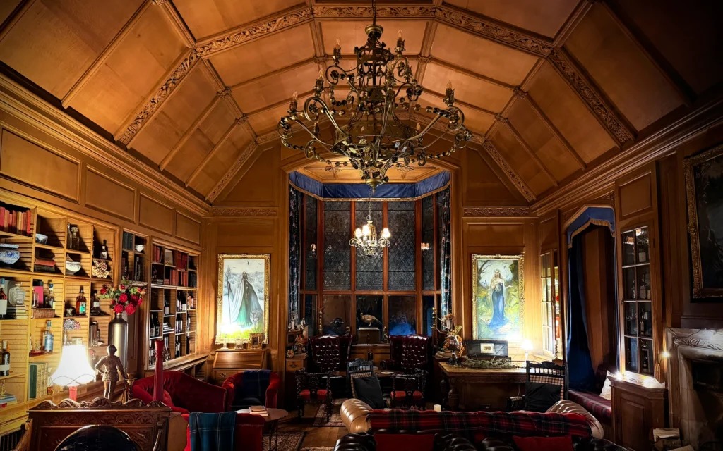 A wood-panelled library with a barrel ceiling and chandelier holds book-lined shelves, stained glass windows and a round table with red armchairs.