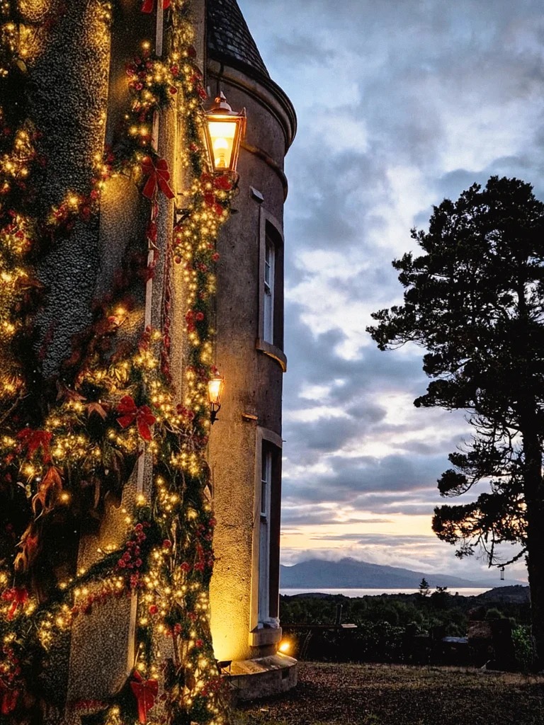 Fairy lights and red-ribboned garland wind up a turreted stone wall at dusk, a lit lantern and hills visible across the water.