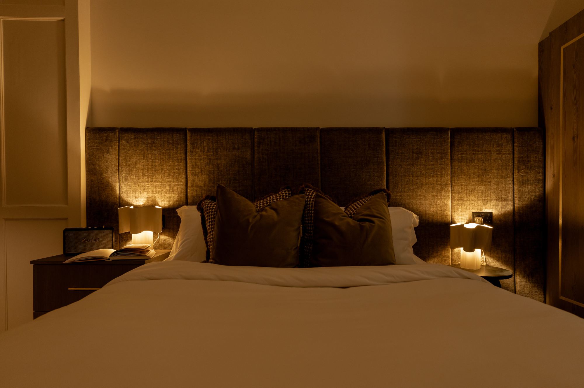 A grey upholstered sofa bed with white and grey pillows sits before a curtained window, a woven pouffe holding a book to the side.