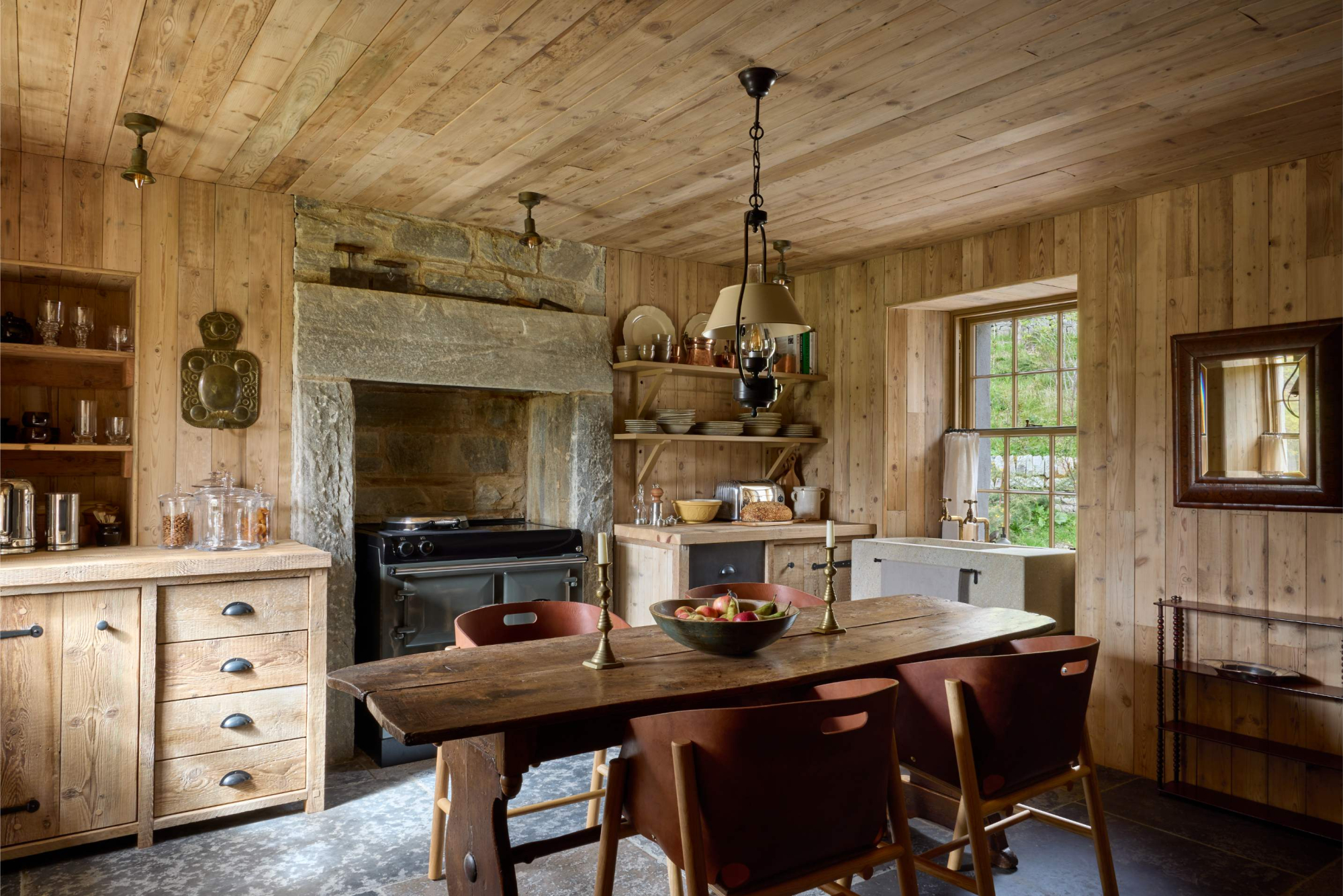 A wooden dining table with leather chairs stands before a stone-arched range and open shelving, a mirror on the panelled wall to the right.