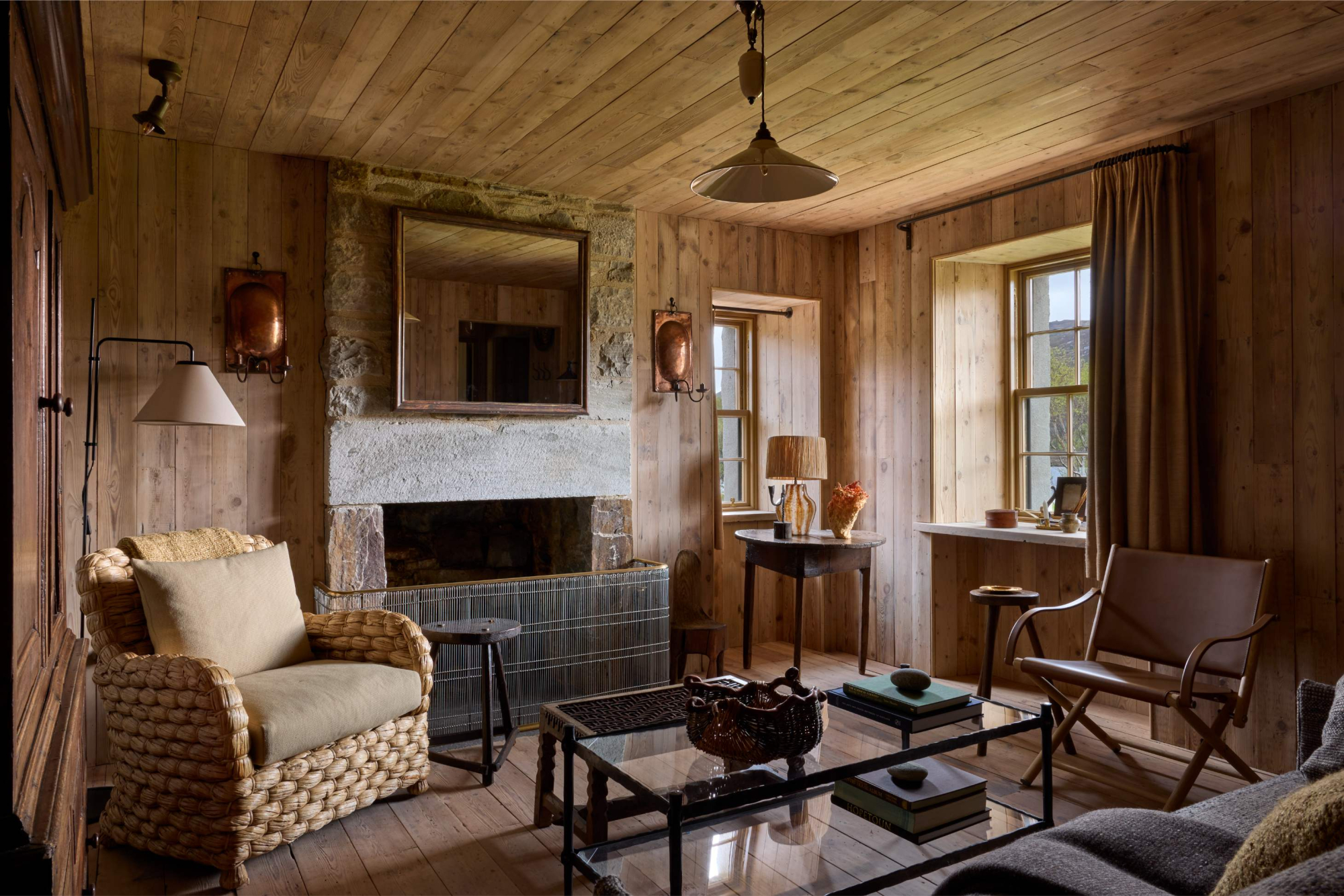 A wicker armchair and leather chair flank a stone fireplace with a mirror above, a glass coffee table in the foreground of a timber-panelled sitting room.