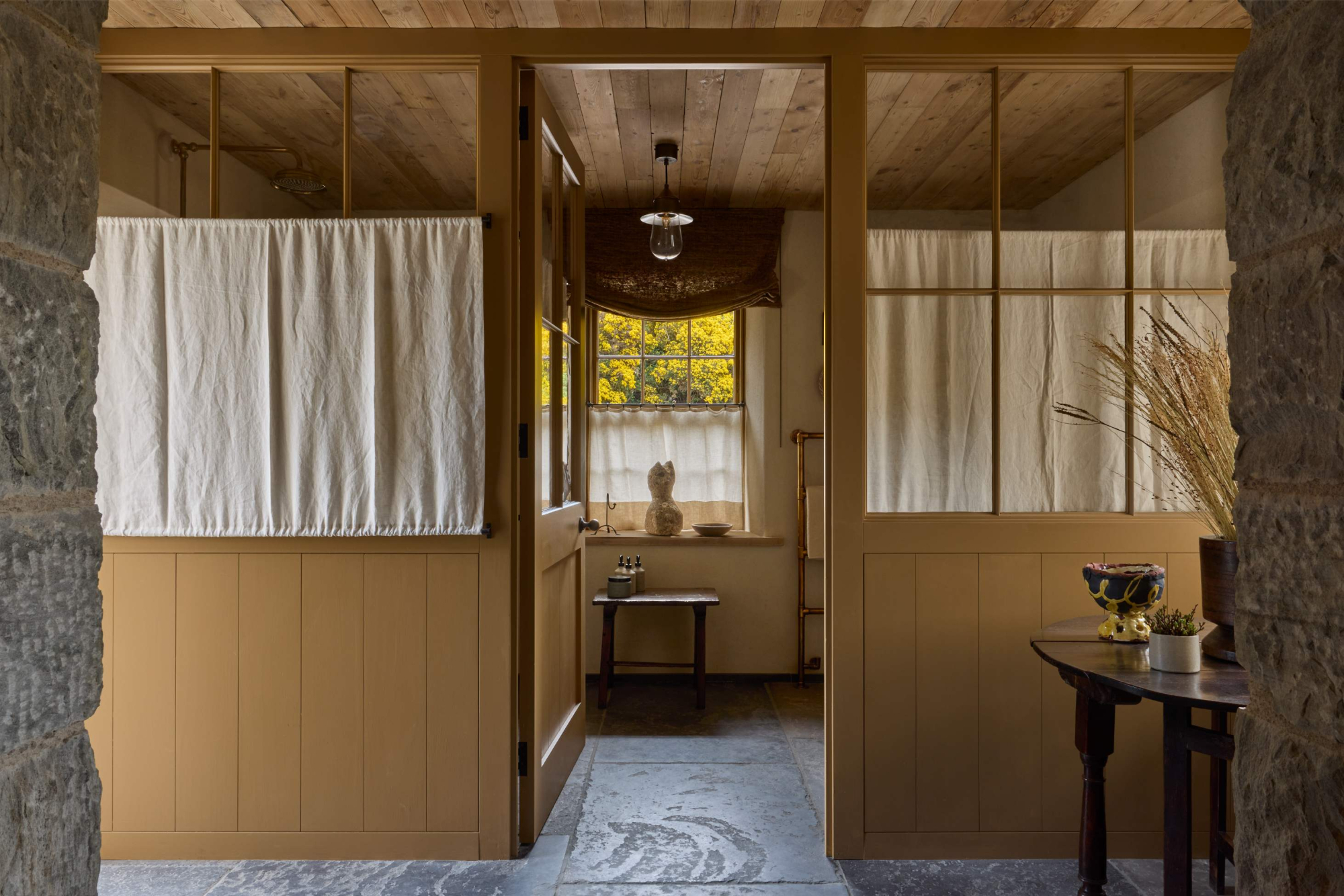 Linen curtains hang either side of a timber-panelled hallway, a small table and stool visible through an open doorway beyond.