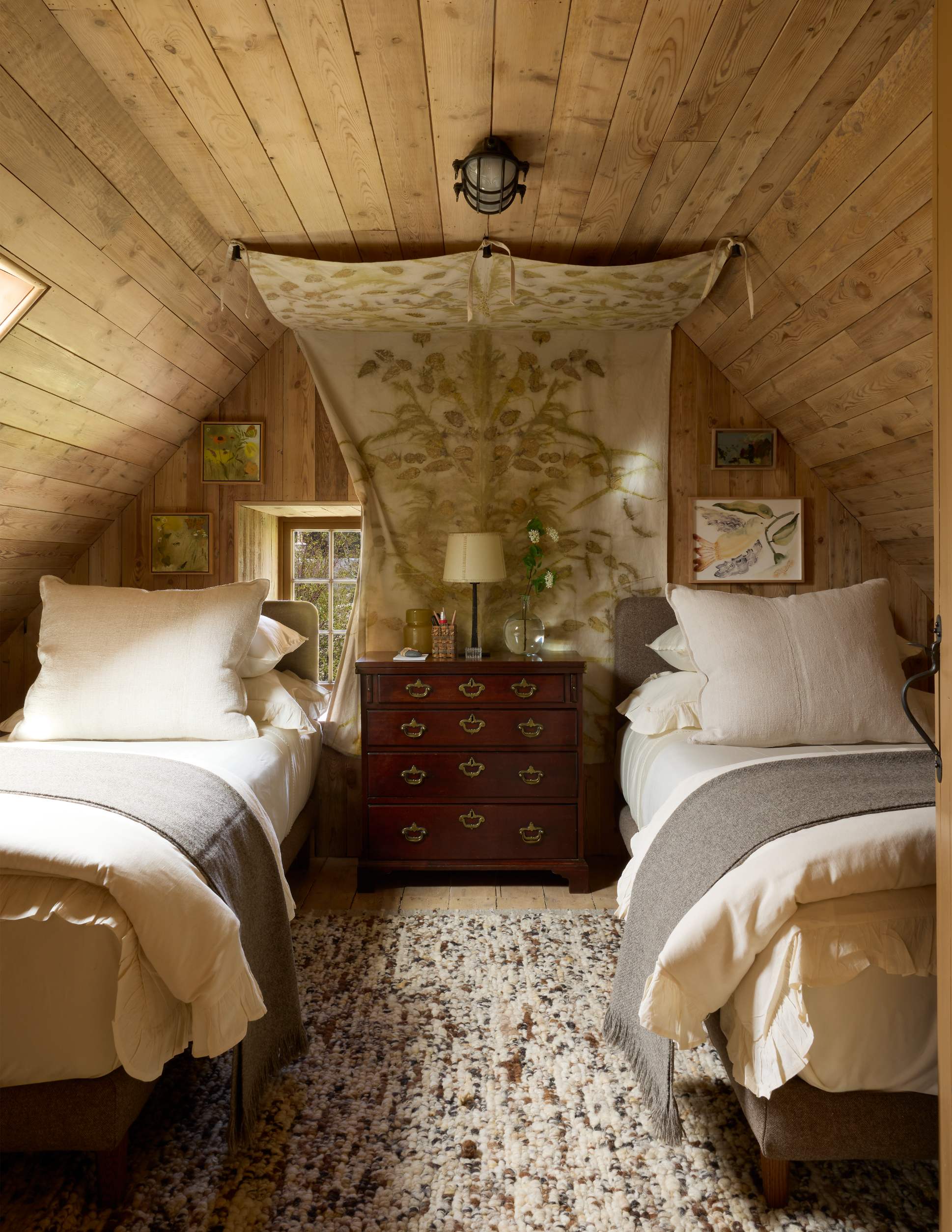 Twin beds with grey throws flank a red chest of drawers beneath a patterned wall hanging, a window and framed prints completing the timber-panelled room.
