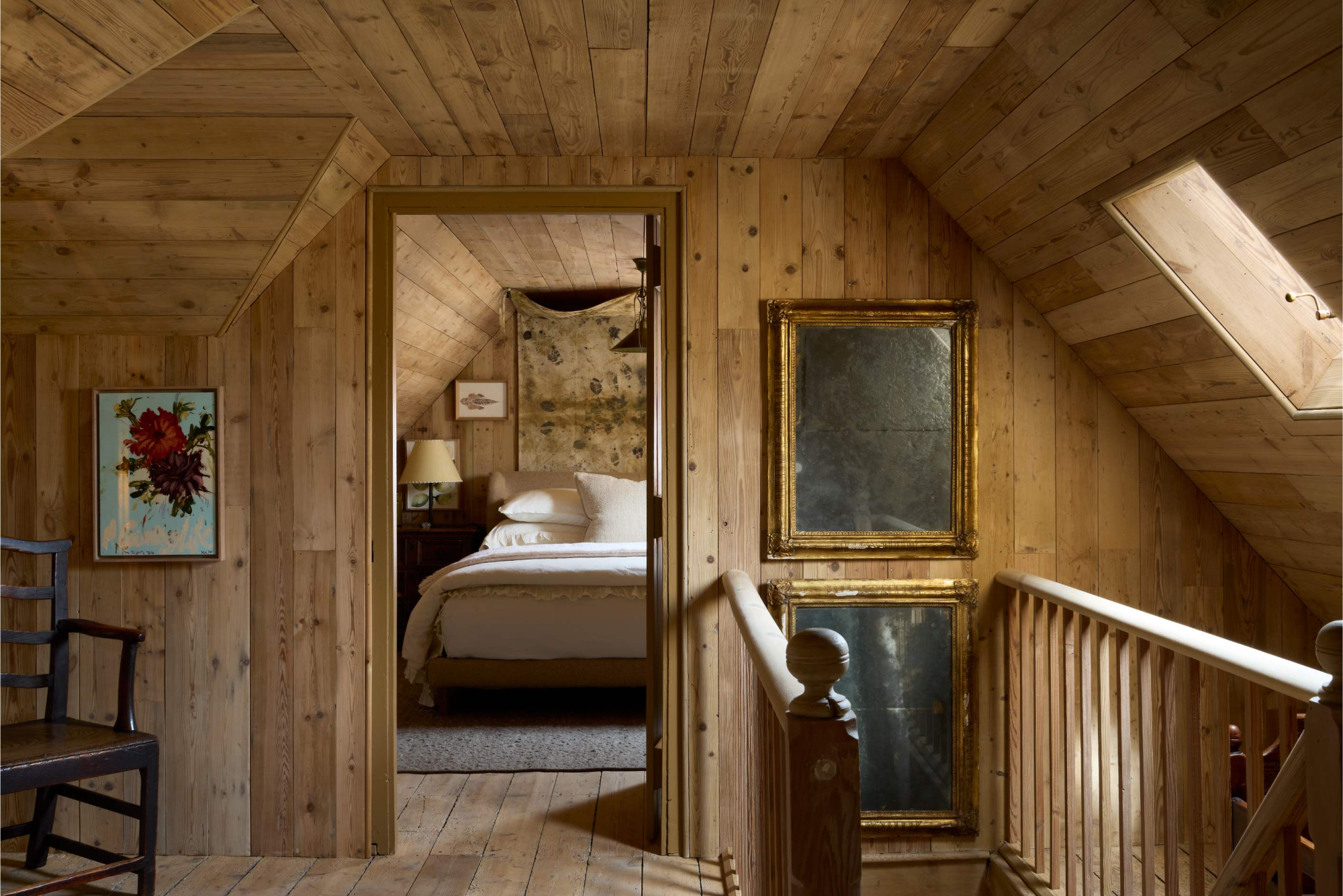 A landing with a wooden banister and gilt-framed mirror looks through an open door onto a bed with white linen and a printed wall hanging.