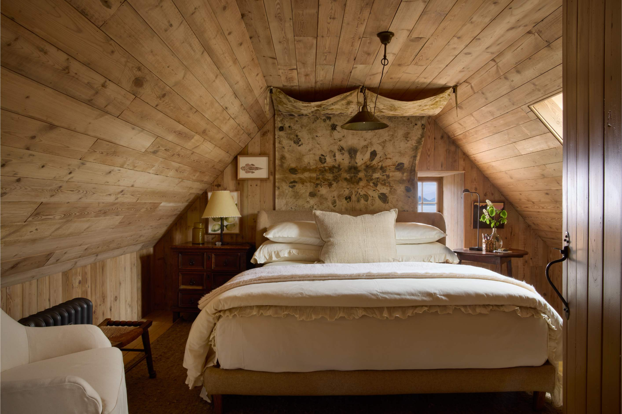 A bed with cream linen sits beneath a stretched canvas hanging from the sloped timber ceiling, a bedside table and vase of flowers to the right.