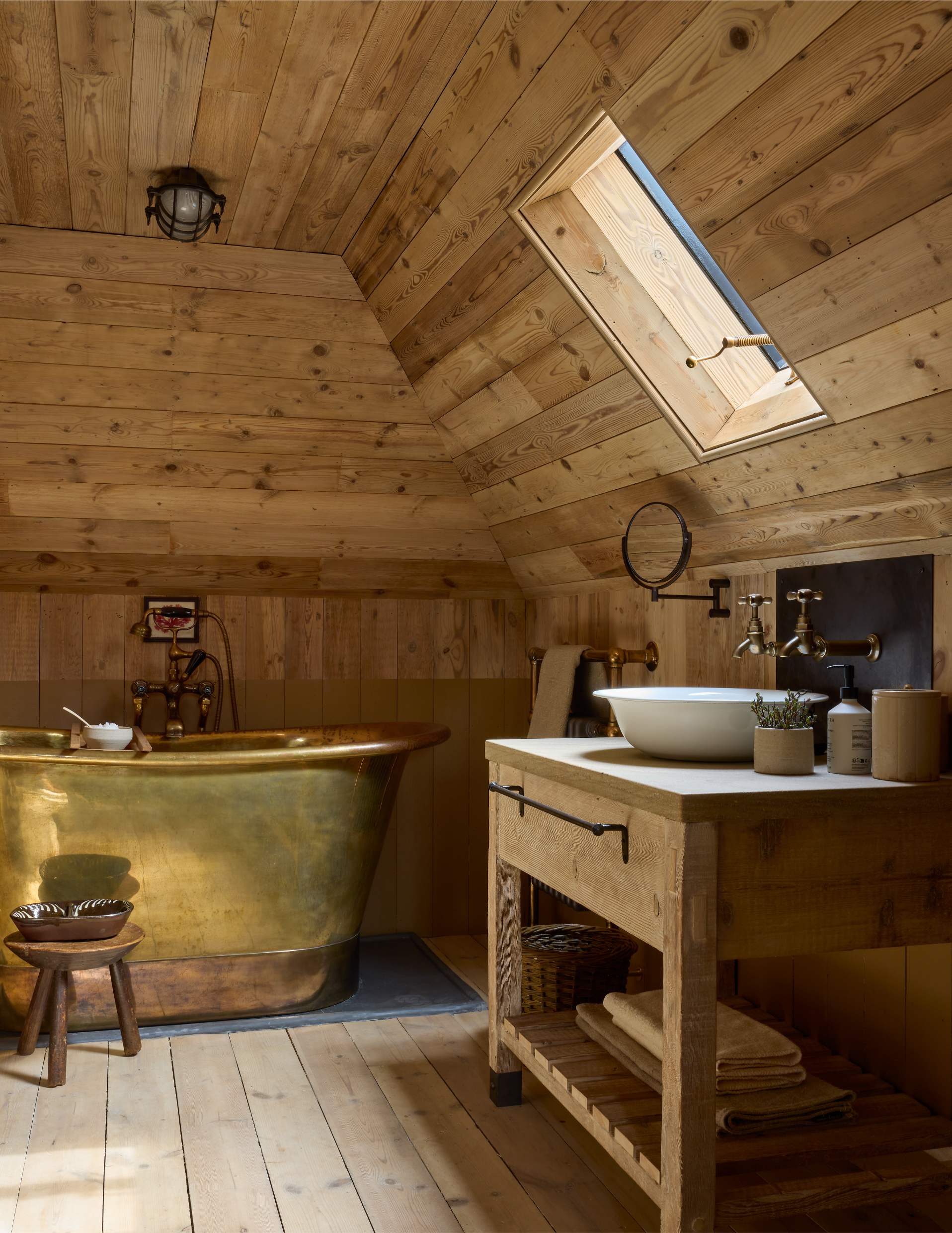 A brass bath sits below a roof window, a wooden vanity with a basin and traditional taps to the right.