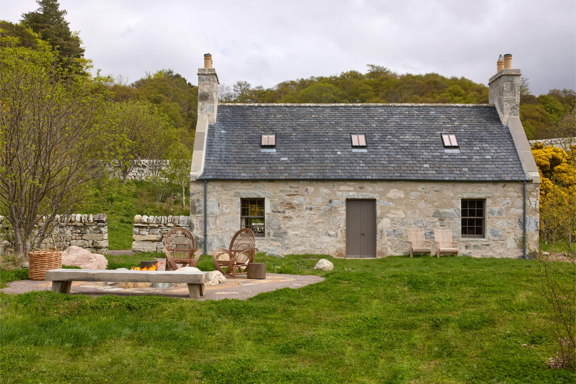 A stone cottage with a slated roof and skylights sits on a lawn, a bench, fire bowl and basket of logs set out on the patio in front.