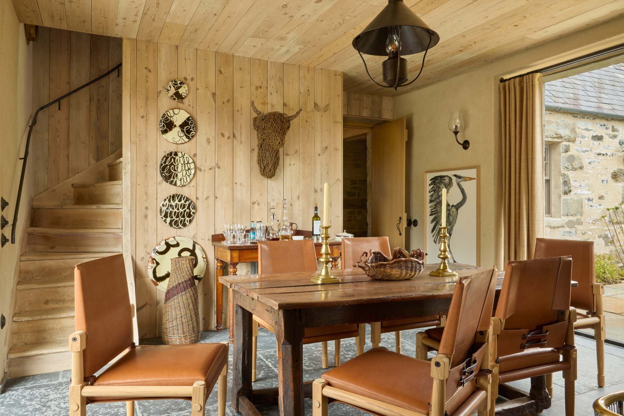 A leather-topped dining table with chairs stands before decorative plates and a woven bull's head on a timber wall, a staircase to the left.