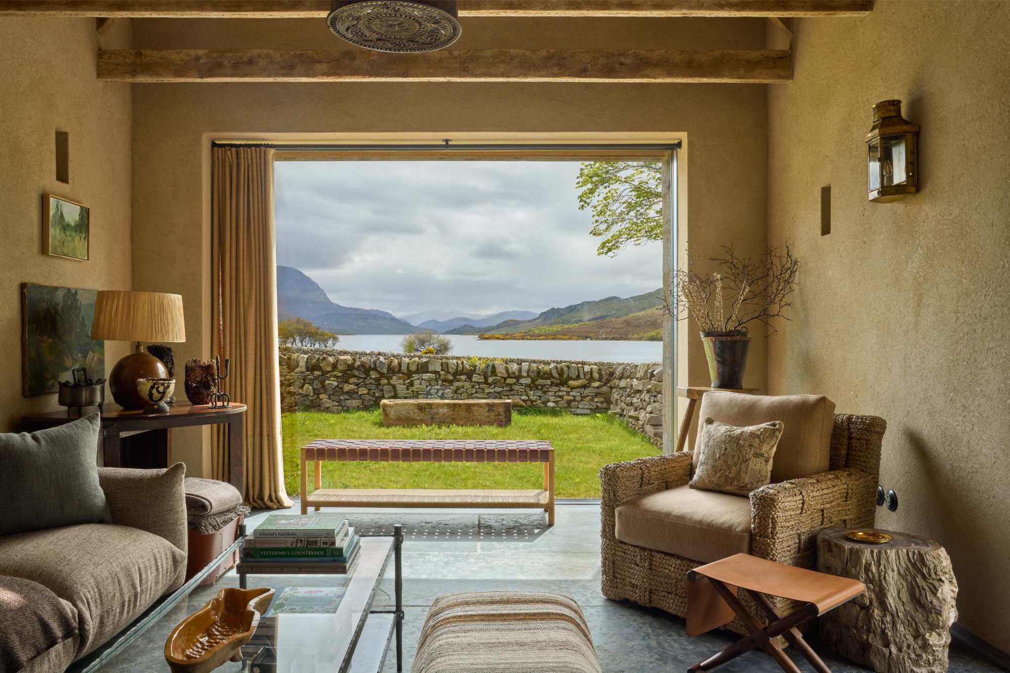 A sofa and wicker armchair sit before open glazed doors looking onto a loch framed by hills, a bench visible on the lawn beyond a dry stone wall.