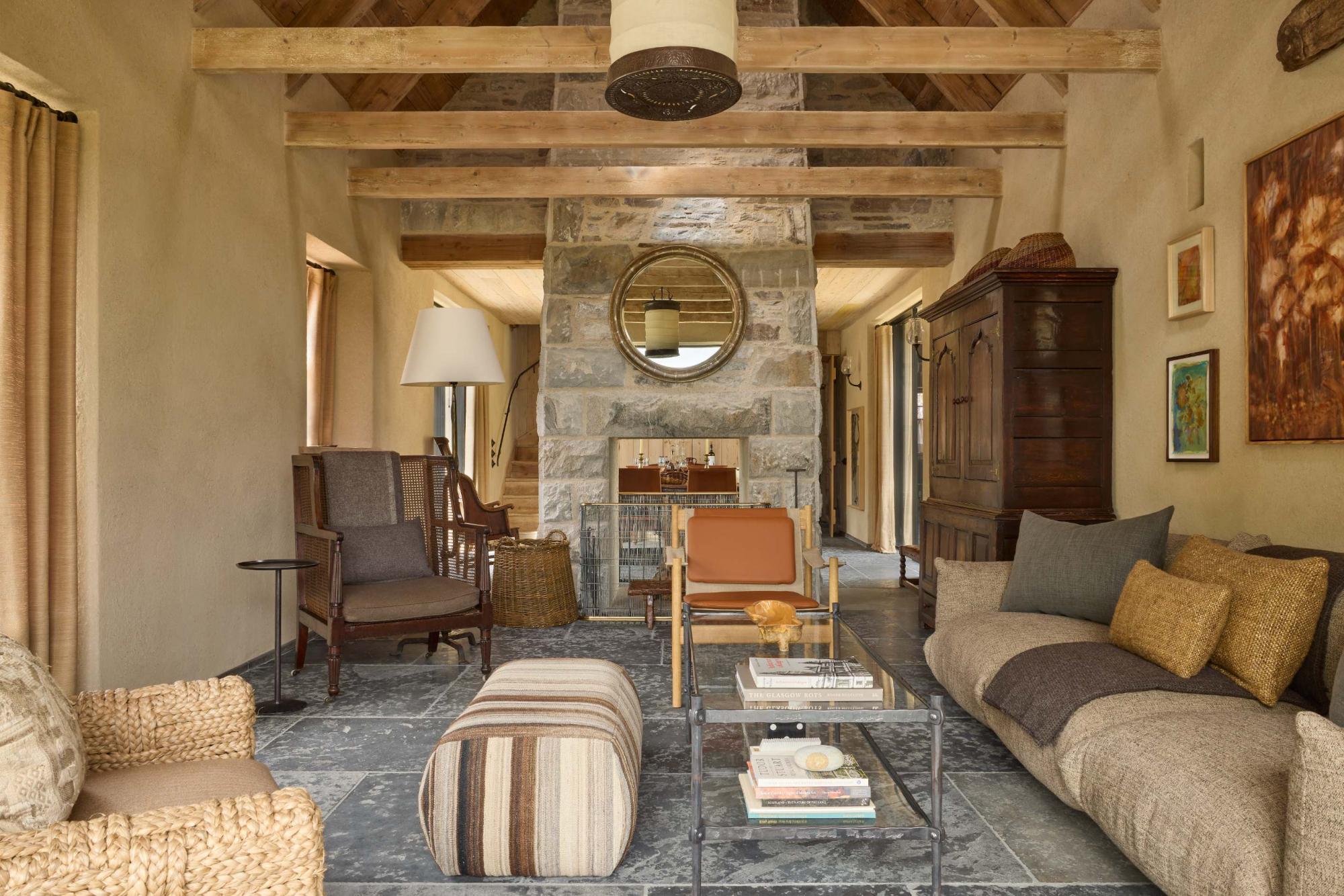A round mirror hangs above a stone fireplace between leather and woven armchairs, a beamed ceiling and sofa completing the sitting room.