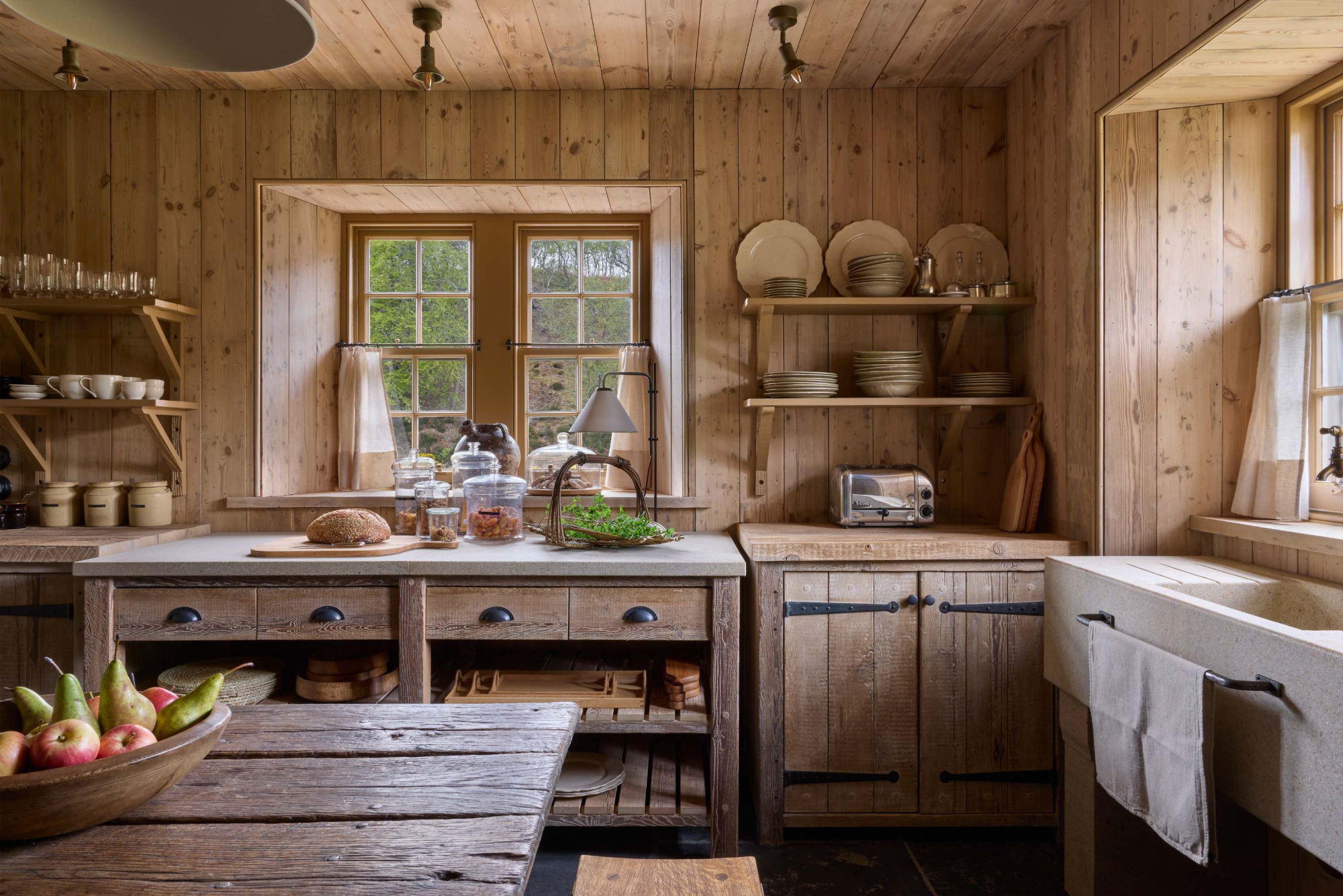 A wooden kitchen island with a bowl of fruit stands before open shelving of plates and a belfast sink, a window seat in the corner.
