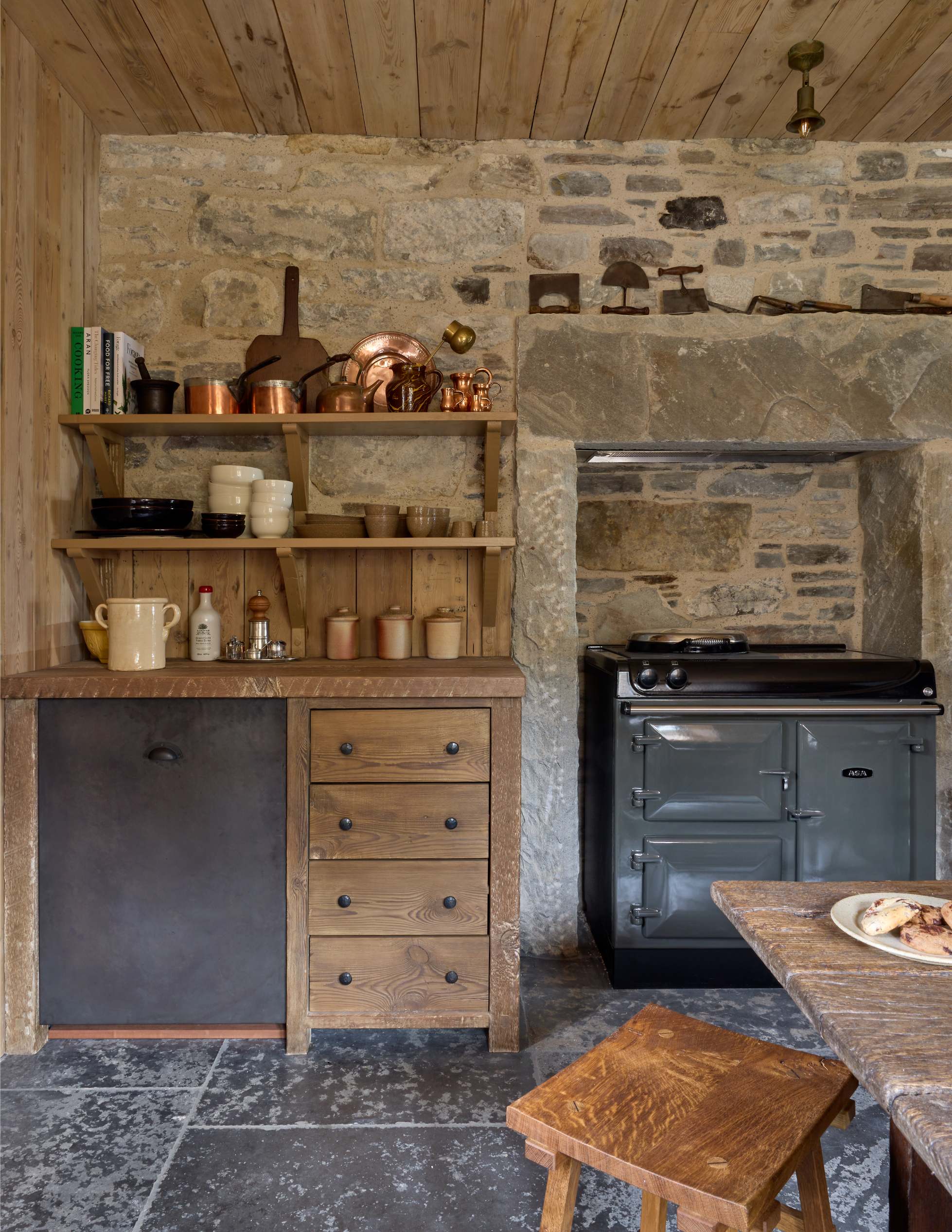 Open shelving holds copper pots and crockery above a dark kitchen unit, a black range cooker set into the stone chimney breast beside it.