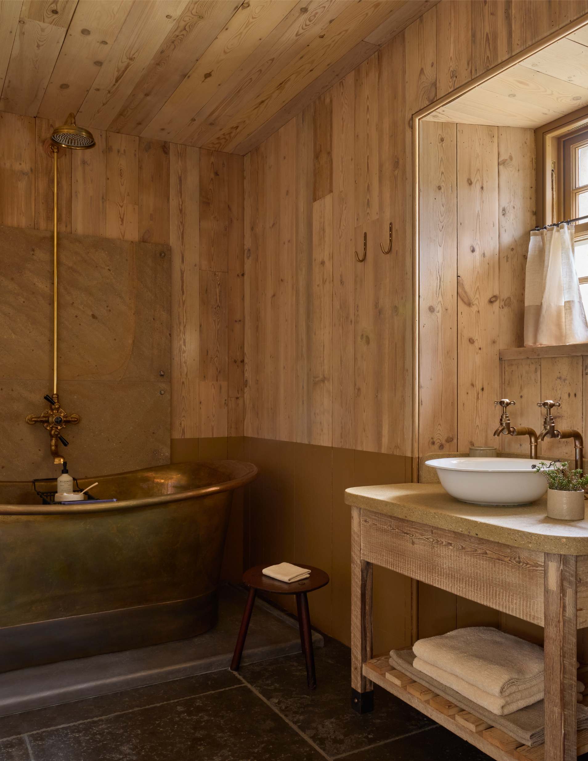 A copper roll-top bath stands below a gold shower head in a timber-panelled bathroom, a countertop basin and folded towels to the right.