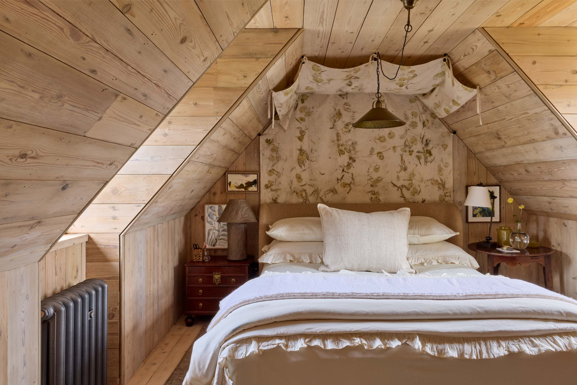 A bed with a floral fabric canopy sits centred beneath a sloped timber ceiling, a dark chest of drawers and lamp to the left.