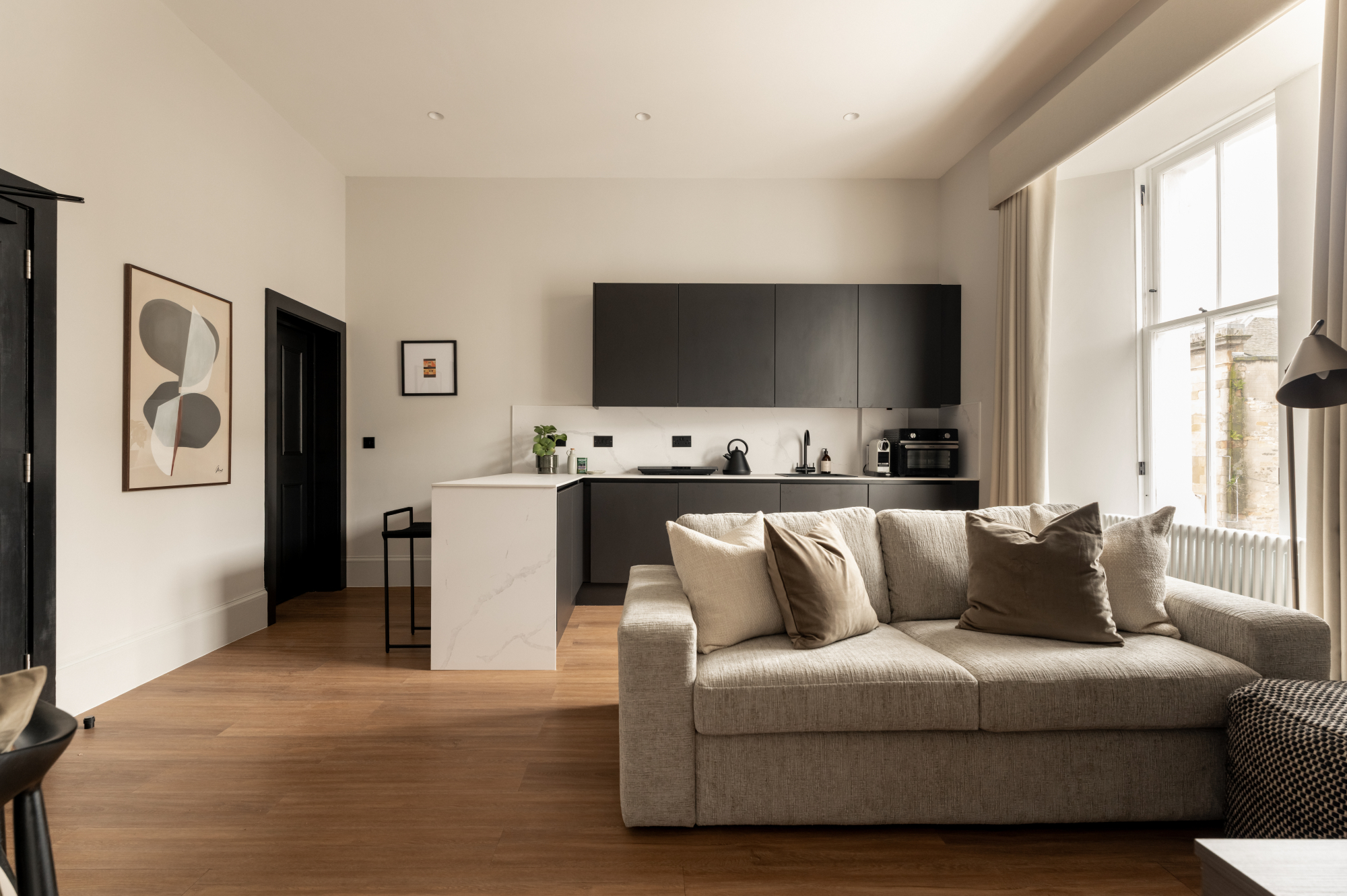 A grey sofa with cushions sits before a black and white kitchen, a framed print on the wall to the left.