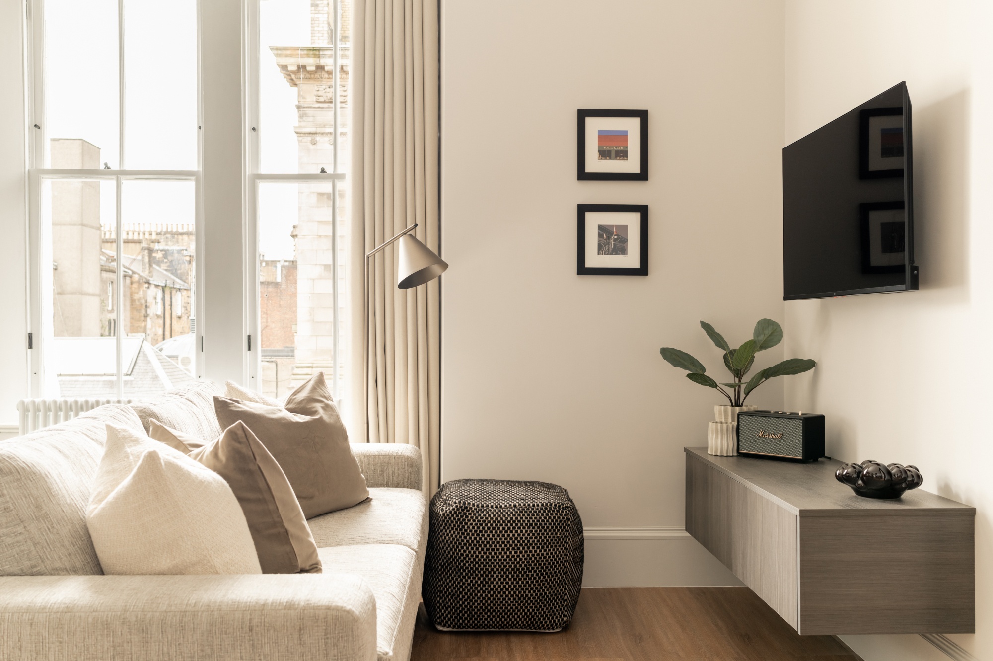 A cream sofa with cushions sits before a mounted television and floating console, framed prints on the wall and a tall window to the left.