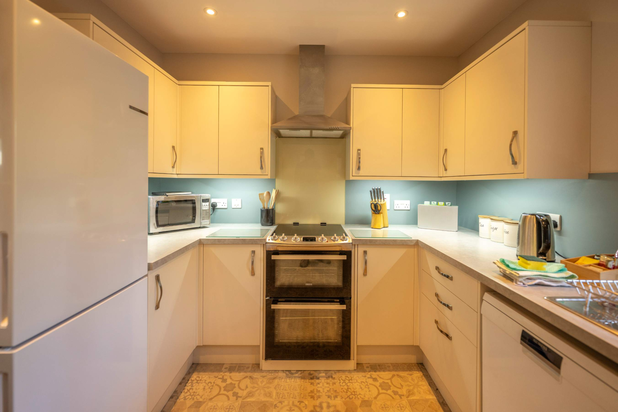 A cream fitted kitchen with a built-in oven and hob, a microwave and kettle on the counter beside patterned tiled flooring.