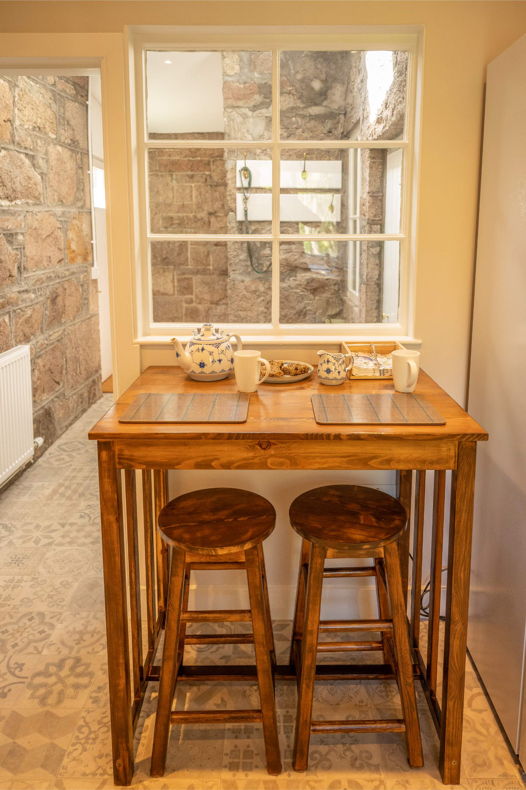 A teapot and mugs are set on a small wooden table with two stools, a stone-walled window behind.