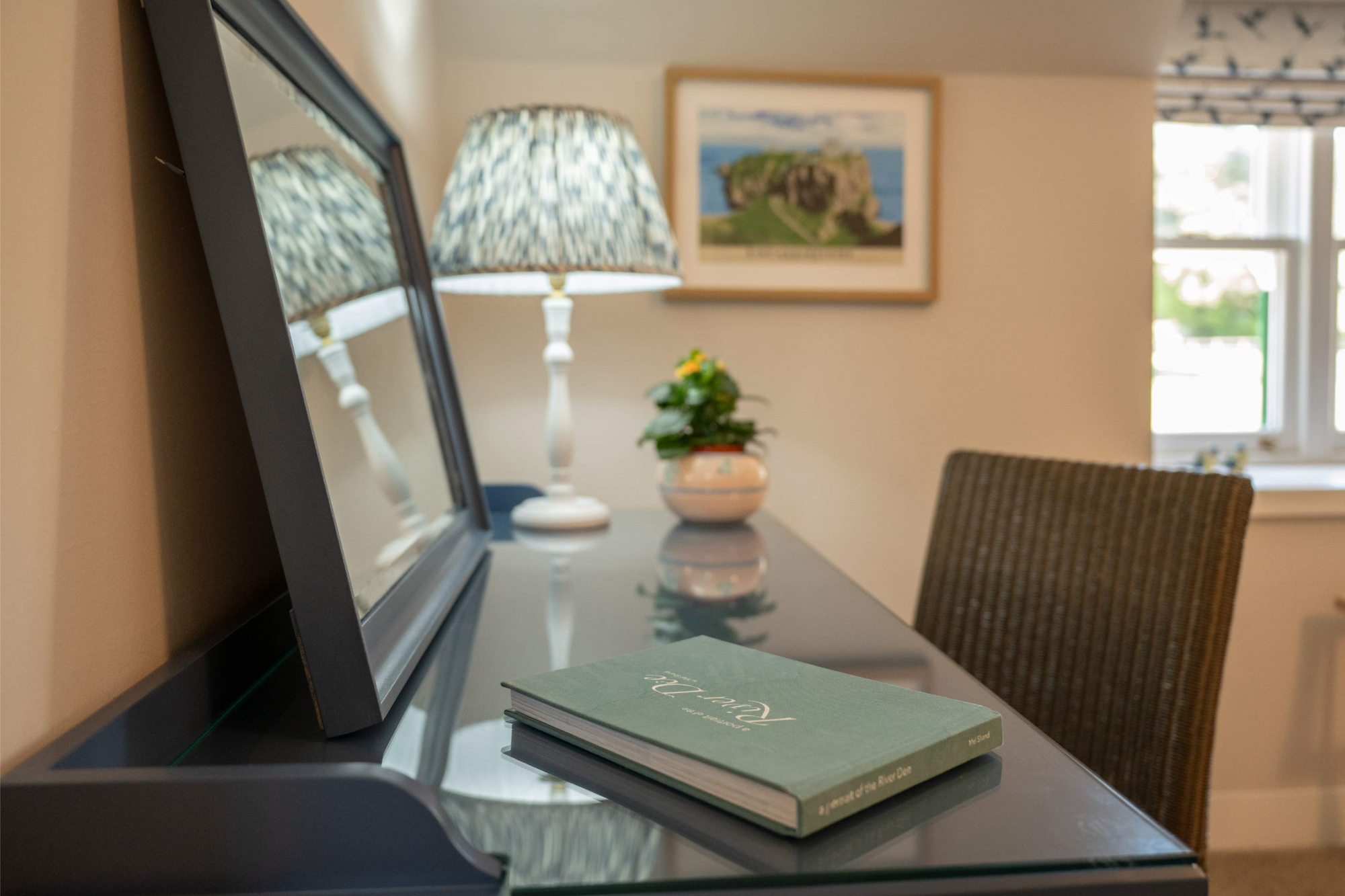 A framed coastal photograph hangs above a desk with a mirror and lamp, a book resting on the glass surface.