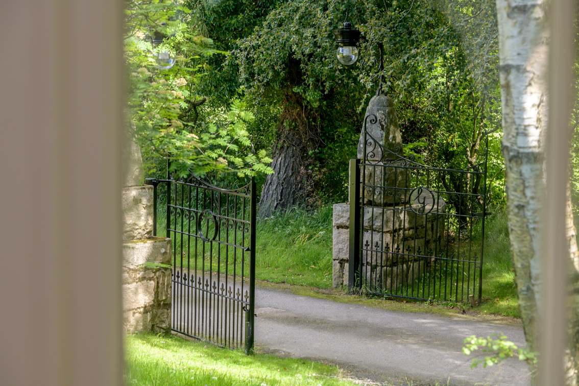 An open wrought iron gate stands before a gravel drive shaded by mature trees, stone gateposts topped with urns either side.