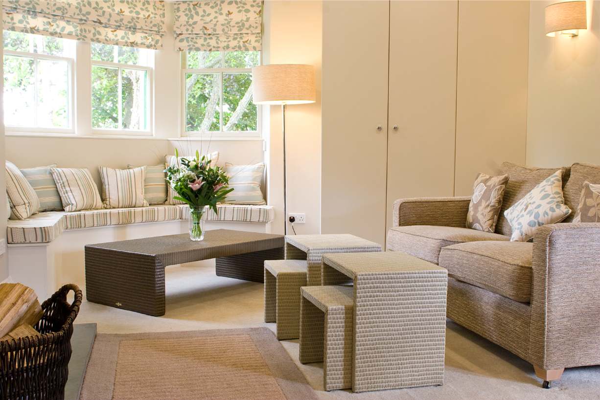 A corner sofa with striped cushions sits below a floral roman blind, a coffee table with a vase of flowers and a woven side table with nested stools completing the room.