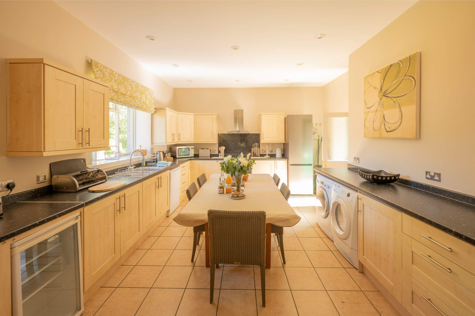 A long kitchen with pale wood units and black worktops, a dining table set with flowers down the centre and a washing machine along the far wall.