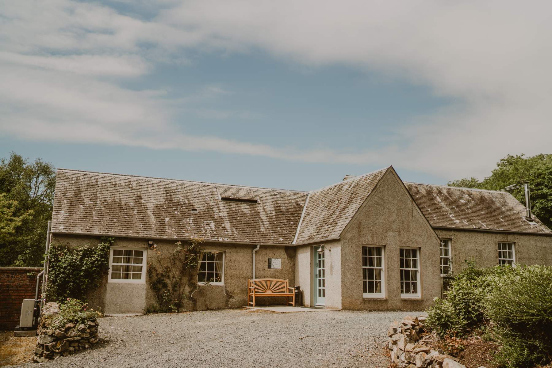 A rendered stone cottage with a gabled porch and a wooden bench sits behind a gravel courtyard, ivy climbing one gable end.