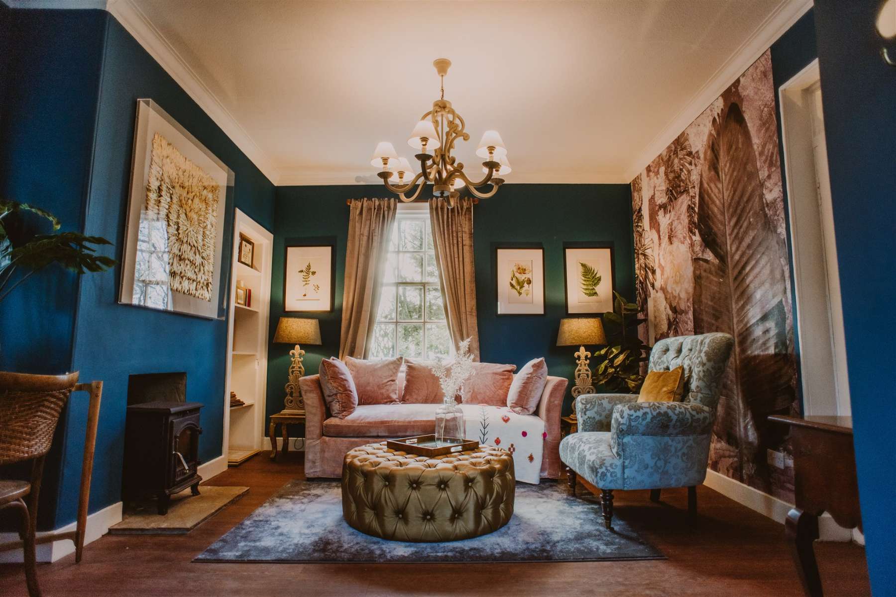 A dusky pink sofa and blue velvet armchair sit before a deep teal wall hung with botanical prints, a wood burning stove and tufted ottoman completing the room.