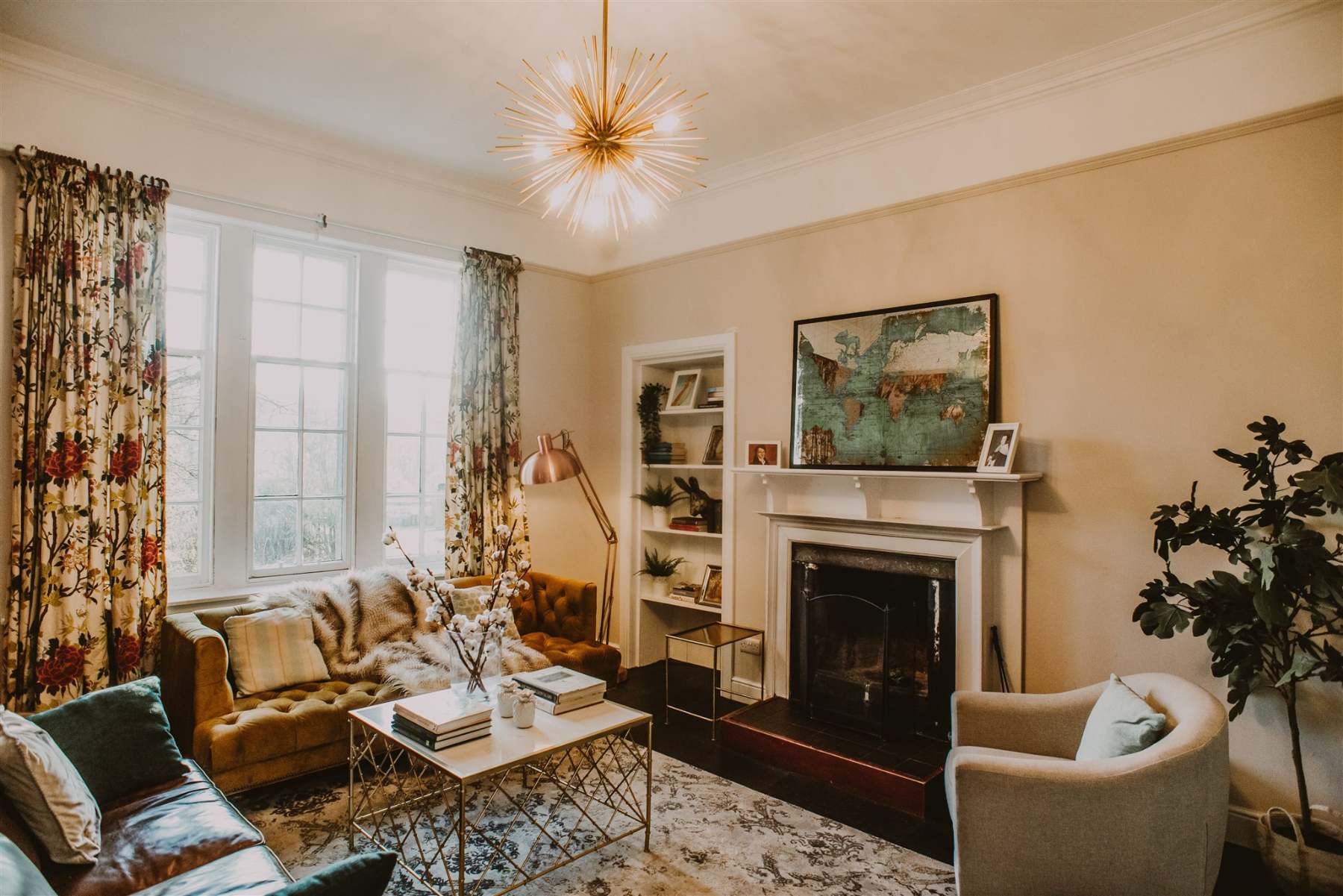 A starburst chandelier hangs above a green sofa and armchair set before a fireplace with a framed painting on the mantel.