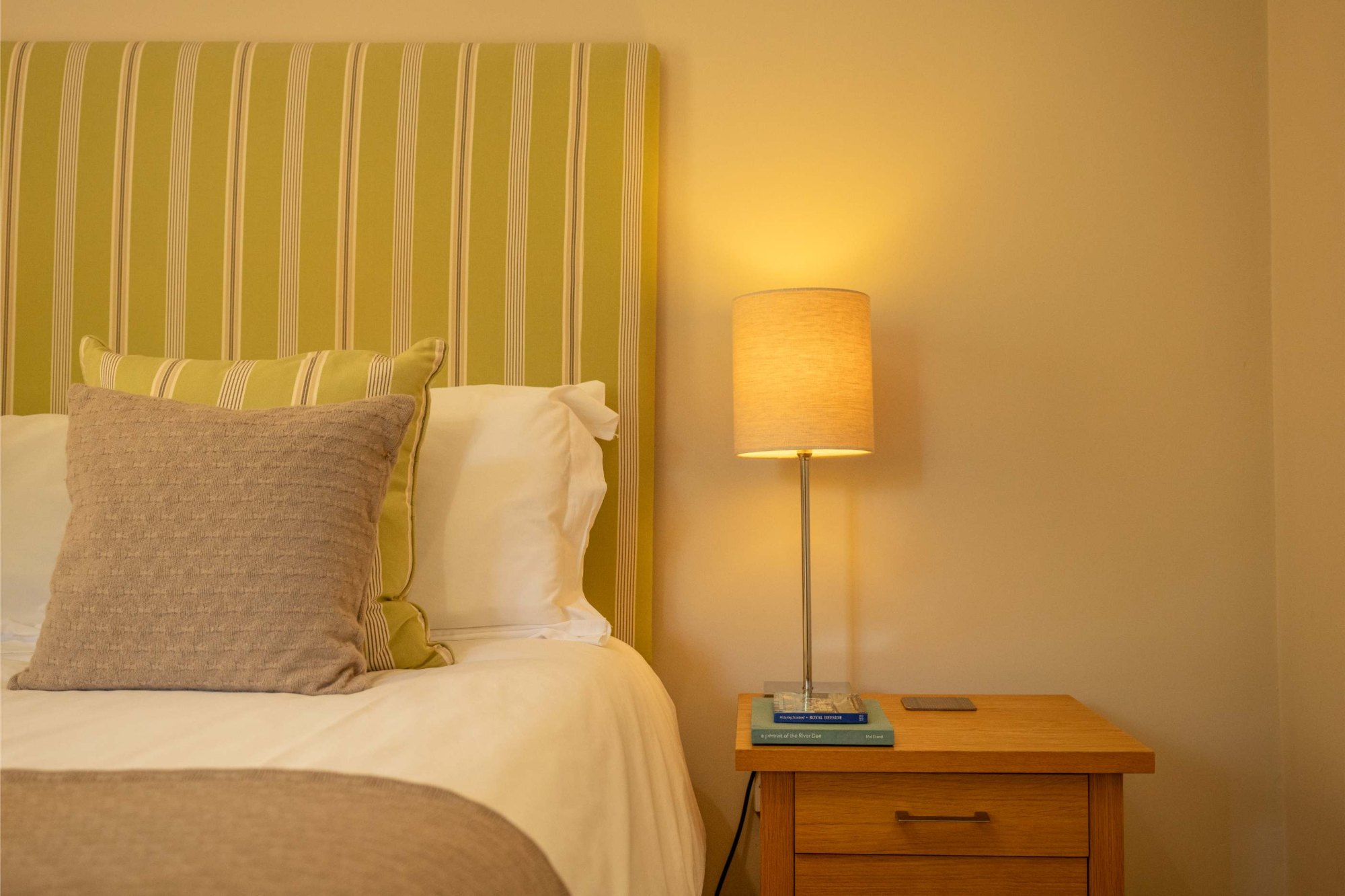 A bed with a green striped headboard and taupe cushions sits beside a wooden nightstand with a lit lamp.