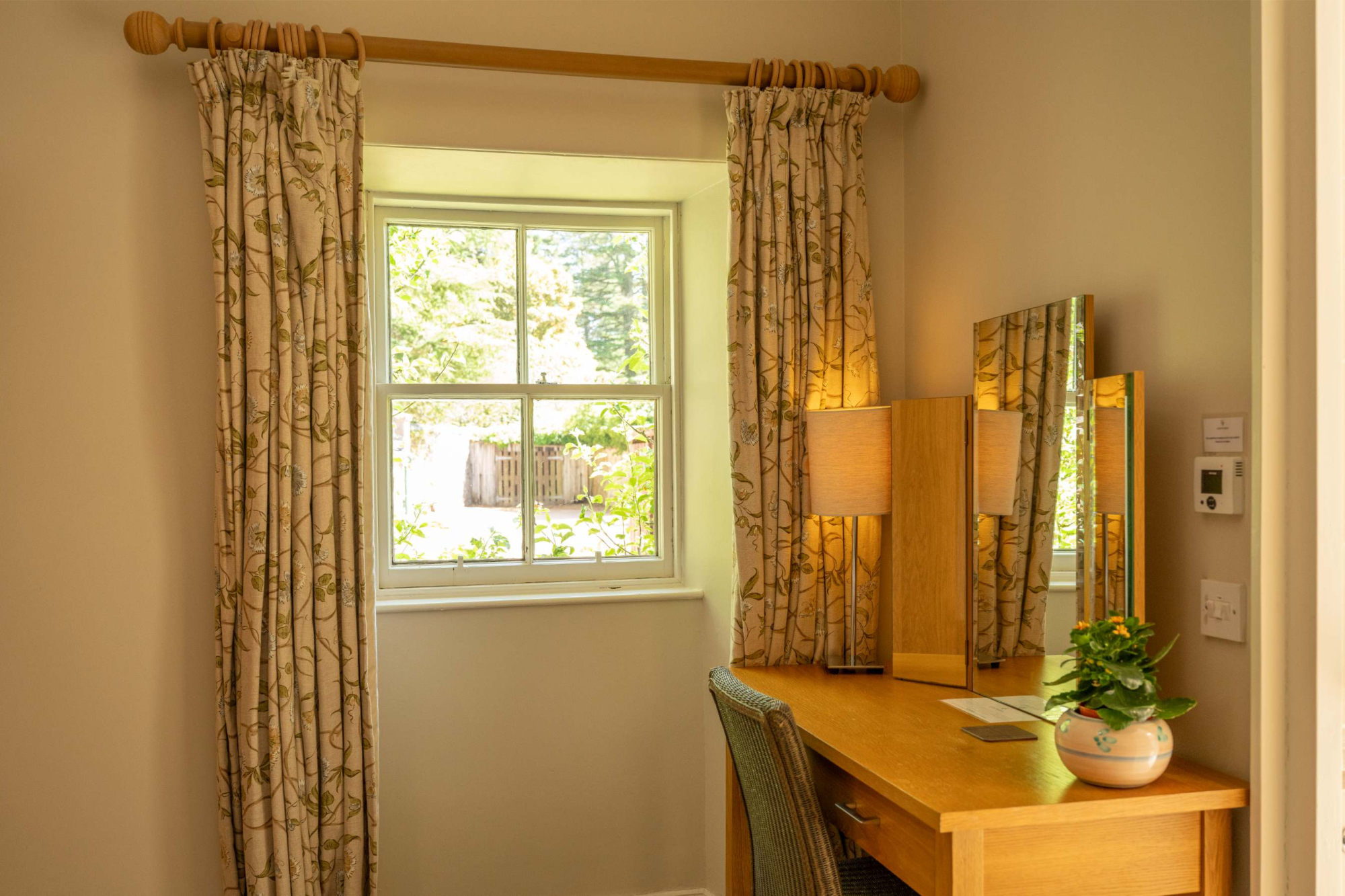 A dressing table with a lit lamp and mirror stands before a curtained window patterned with birch trees, a green chair tucked beneath.