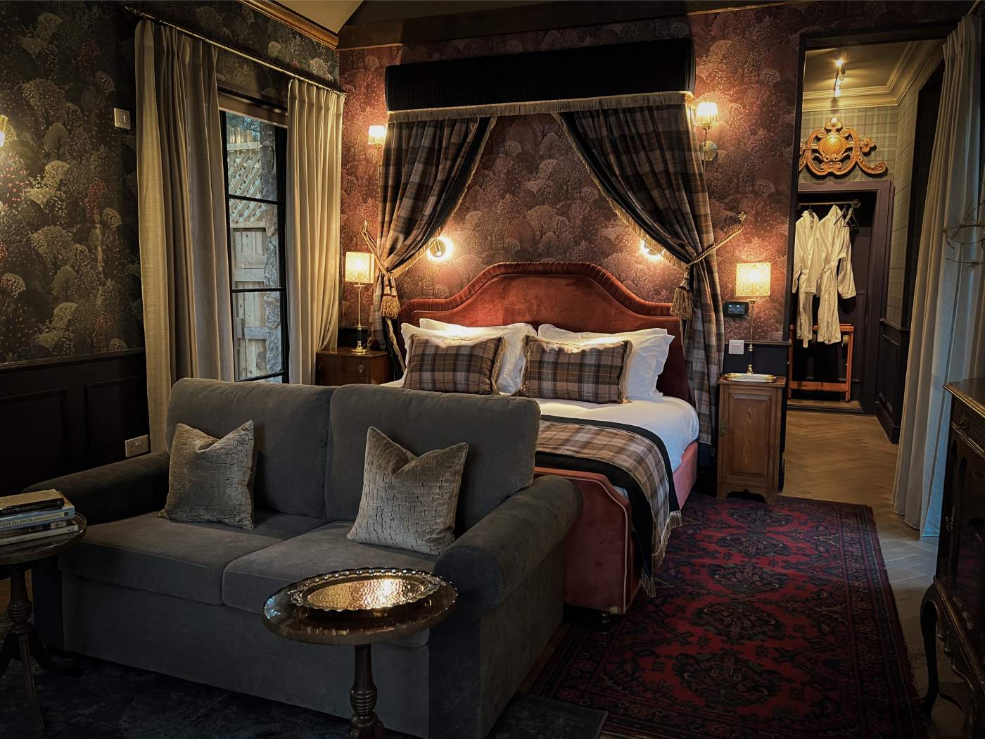 A grey sofa with cushions sits before a bed with a red upholstered headboard and tartan cushions, a doorway showing a robe hanging beyond.