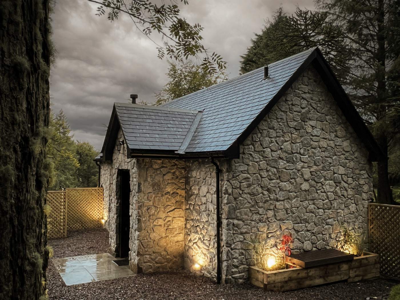A stone bothy with a slated roof and small gabled porch sits among trees, outdoor lighting and a bench beside potted flowers.