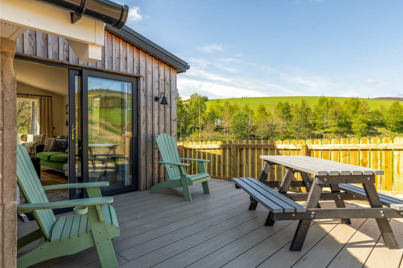 Two green armchairs sit on a wooden deck beside a picnic bench, glazed doors opening onto a sofa inside and fields beyond the fence.
