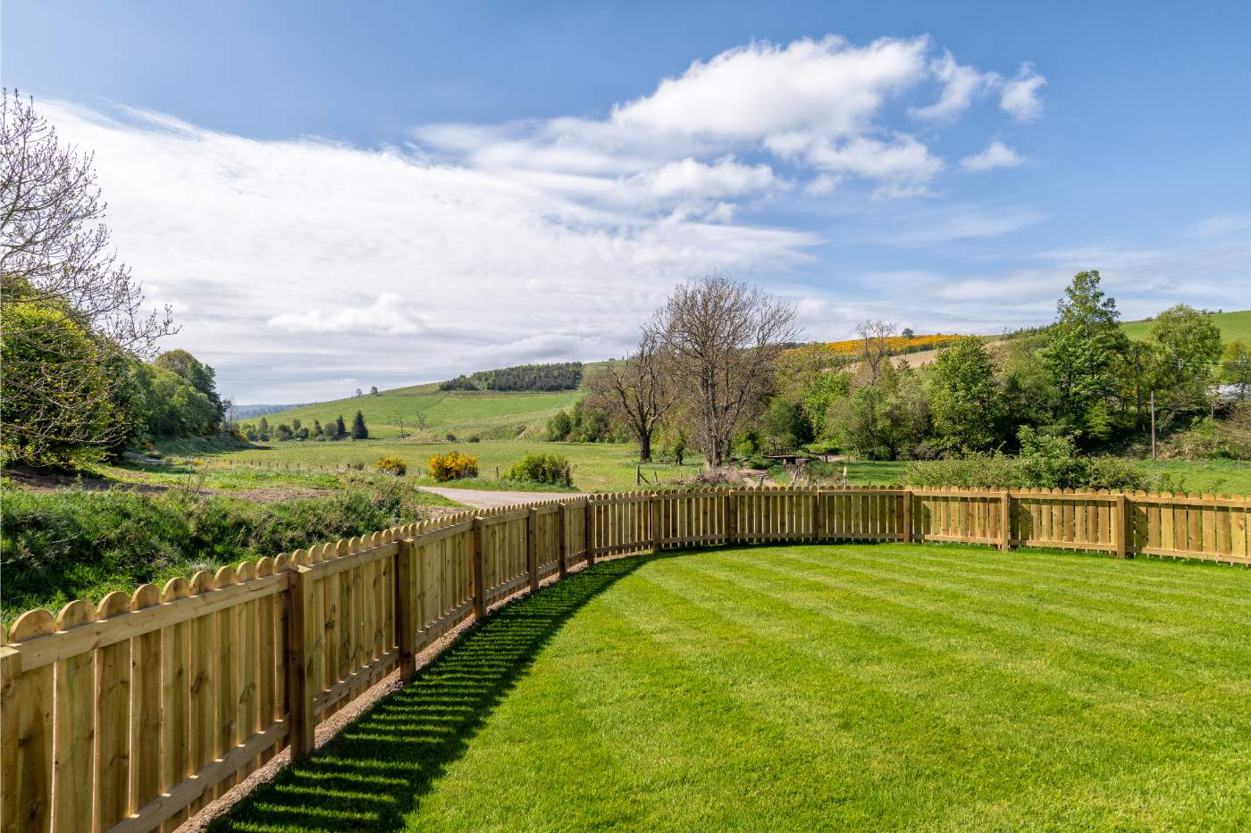 A lawn edged by a curved timber picket fence looks out over rolling green hills and scattered trees.