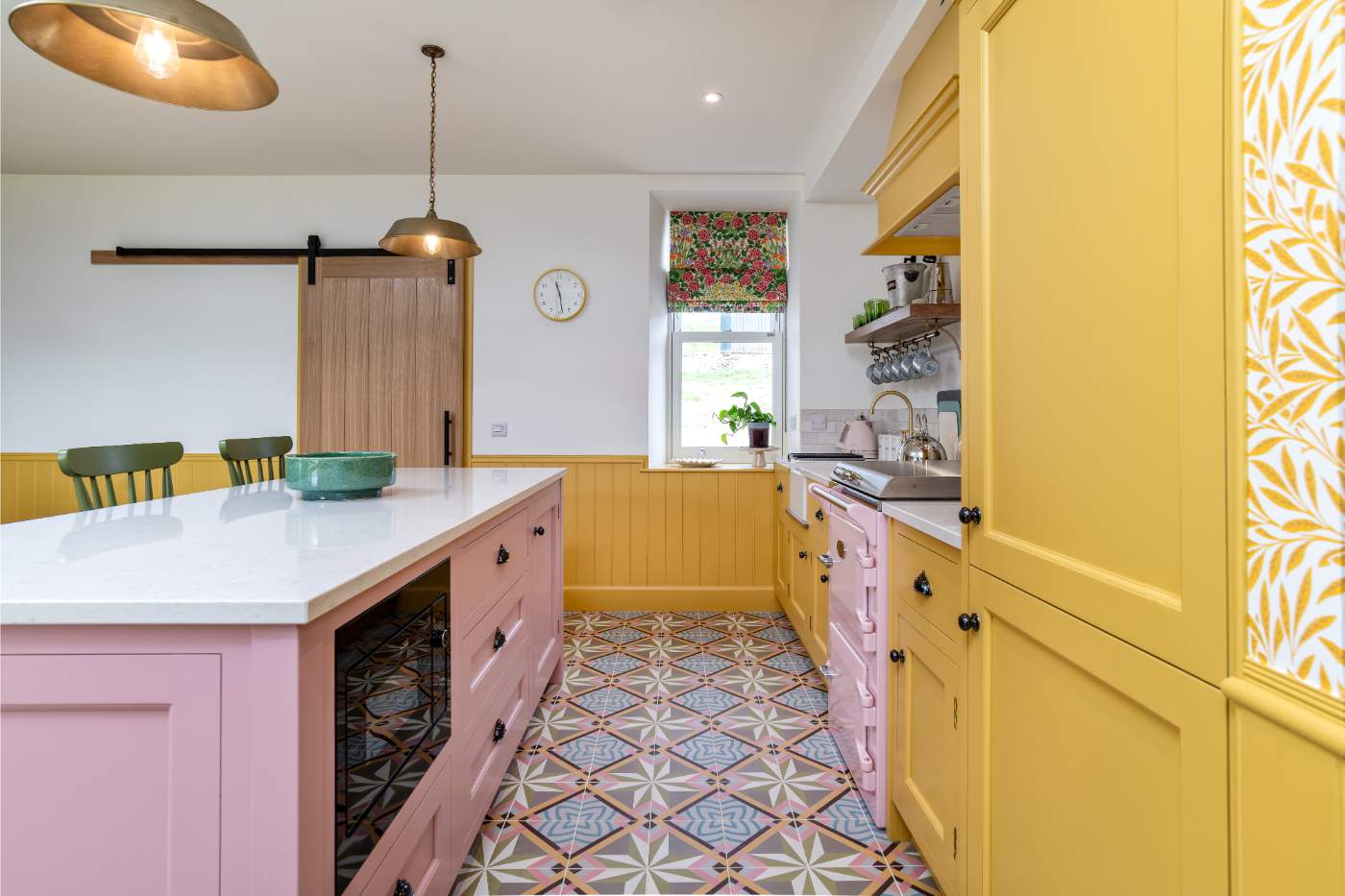 A kitchen with mustard yellow cabinetry and a pink island, a barn door and patterned tiled floor beneath pendant lights.