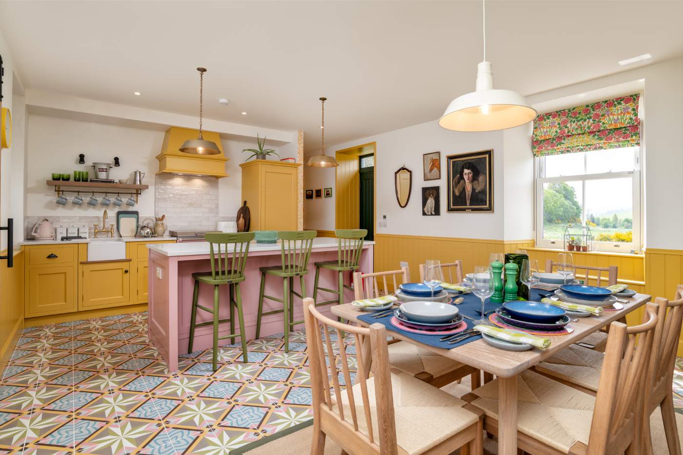 A dining table set for a meal stands before a kitchen with a pink island and yellow units on a patterned tiled floor.