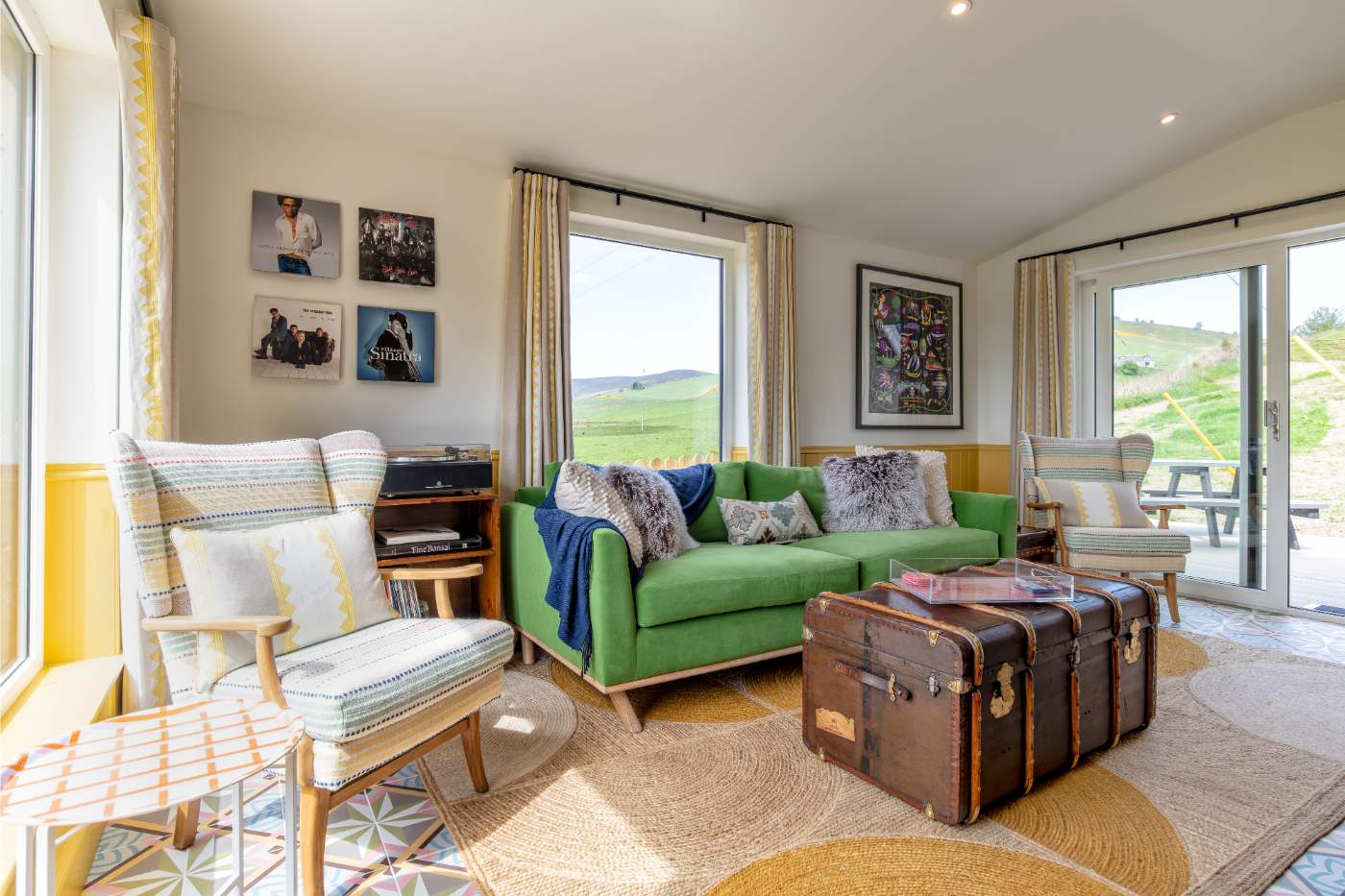A green sofa with a tartan throw sits before a window looking onto fields, armchairs and a trunk coffee table completing the room.