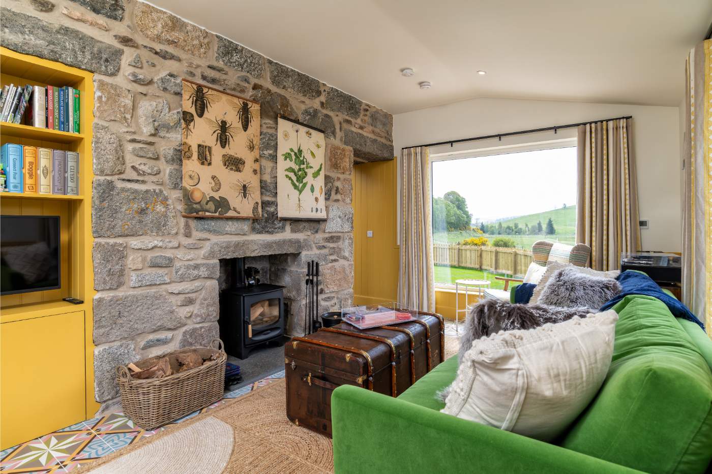 A green sofa piled with cushions sits before a stone fireplace hung with insect and botanical prints, glazed doors opening onto a lawn.