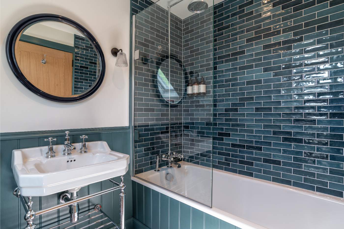 A dark green tiled bathroom with a bath and shower screen, a round mirror above a pedestal basin with traditional taps.