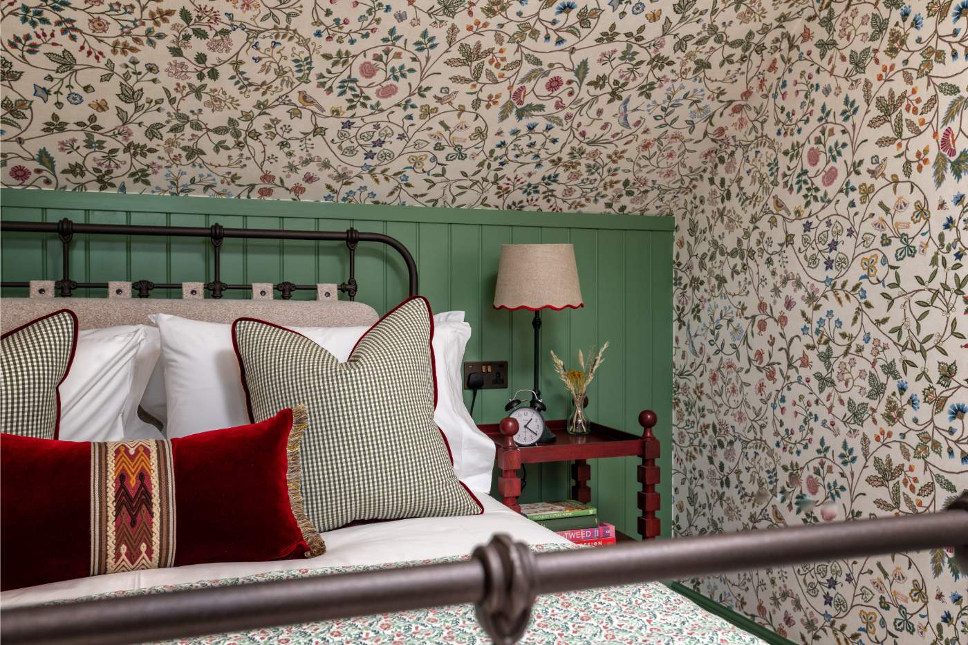A wrought iron bed piled with red and check cushions sits against green panelling, floral wallpaper covering the sloped ceiling and a red side table with a lamp.