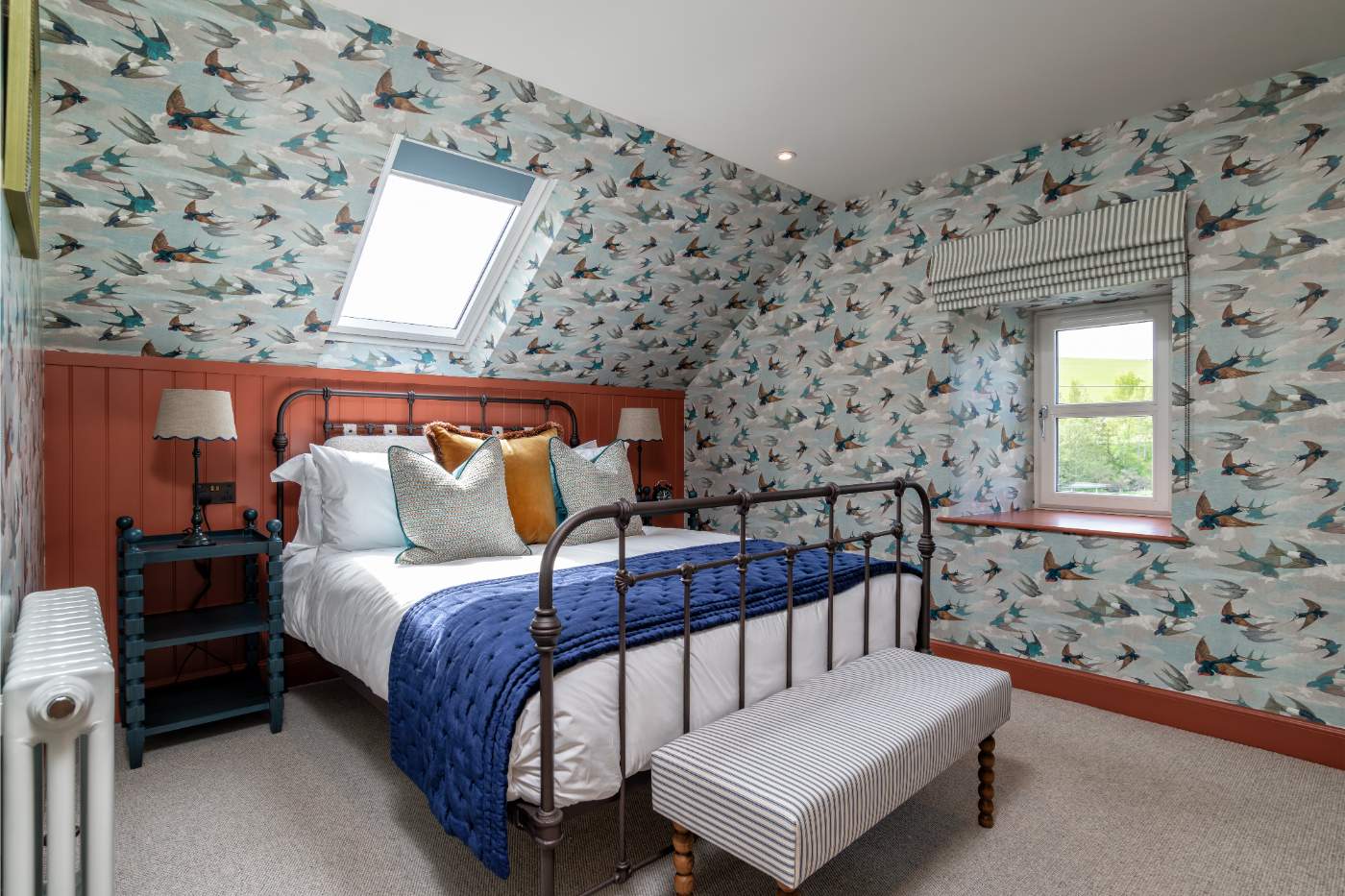 A wrought iron bed with a blue throw sits against terracotta panelling, bird-print wallpaper covering the sloped ceiling above.
